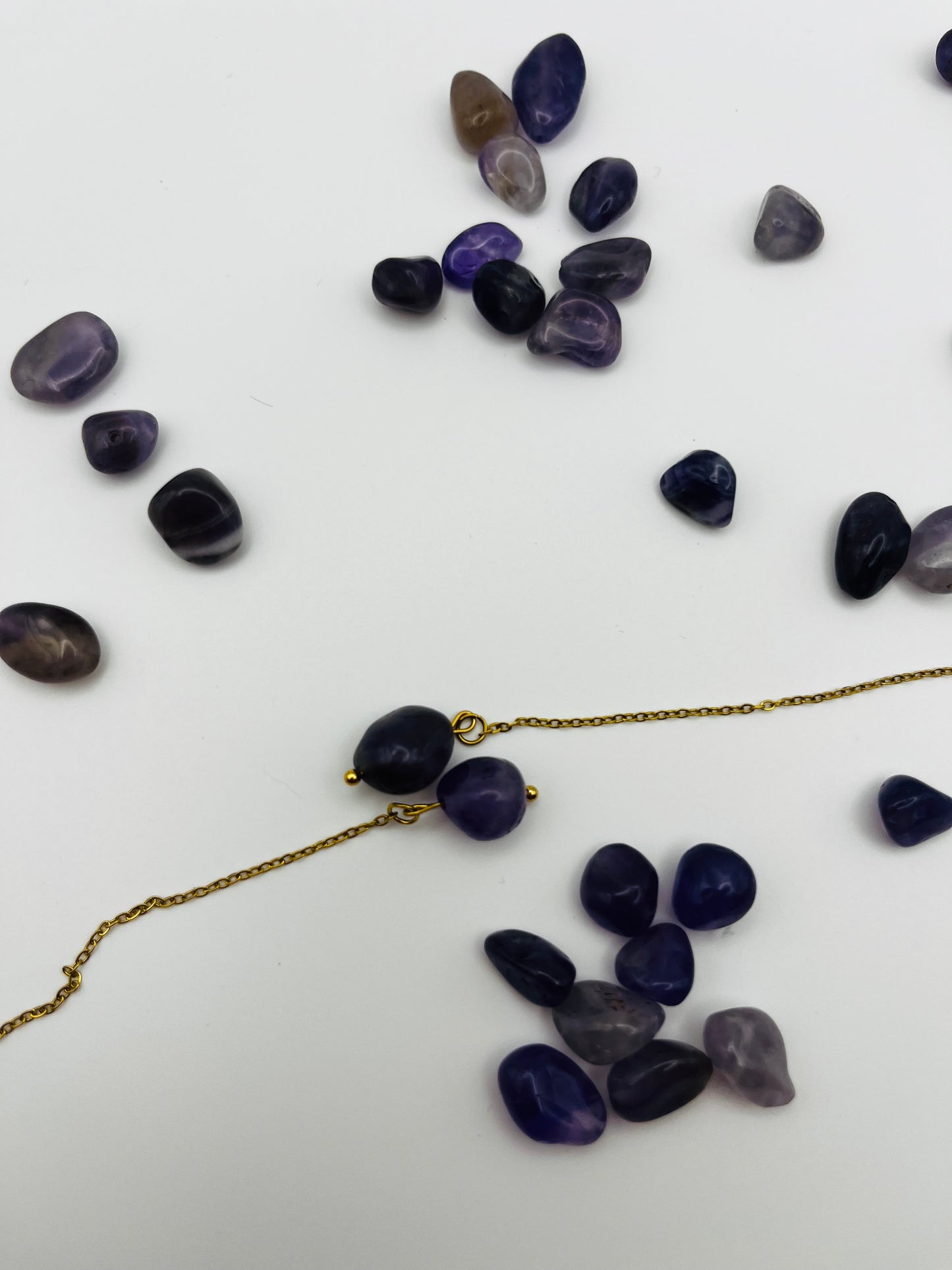 Pendientes Délica – detalle en horizontal del gancho threader dorado, cadena dorada fina y dos cuentas ovaladas de amatista morada oscura natural con bolita dorada, con cuentas de amatista morada de distintos tonos sueltas esparcidas, sobre fondo blanco, hechos a mano en Murcia
