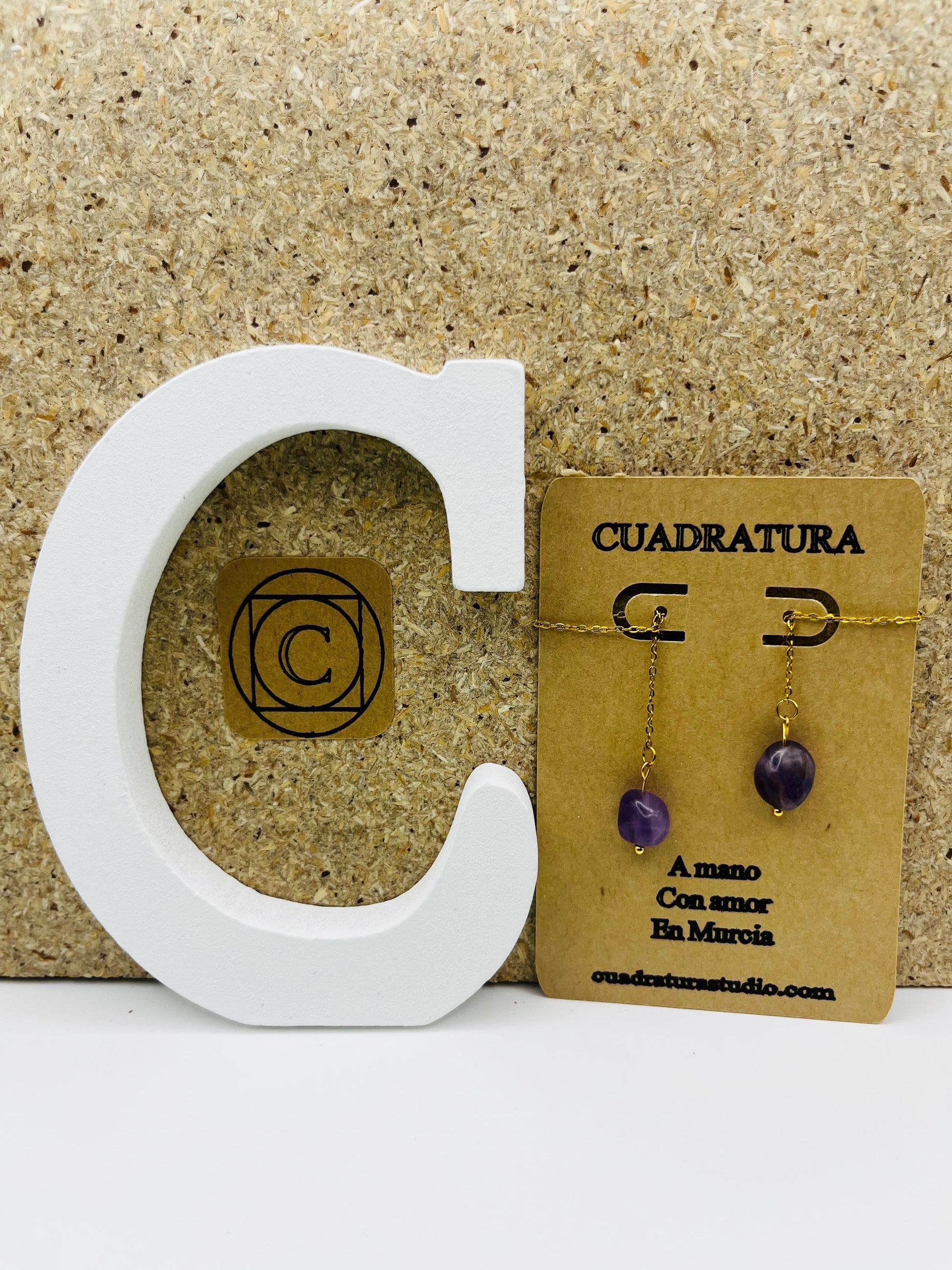 Pendientes Délica – pendientes simétricos con gancho dorado, cadena dorada fina y cuenta ovalada de amatista morada natural, en tarjeta Cuadratura, sobre letra C blanca y fondo de corcho, hechos a mano en Murcia