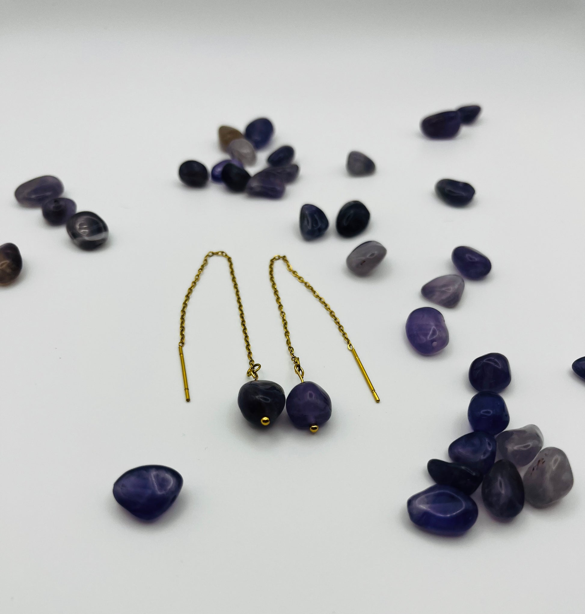 Pendientes Délica – pendientes simétricos con gancho threader dorado, cadena dorada fina y cuenta ovalada de amatista morada oscura natural, con cuentas de amatista morada de distintos tonos sueltas esparcidas, sobre fondo blanco, hechos a mano en Murcia