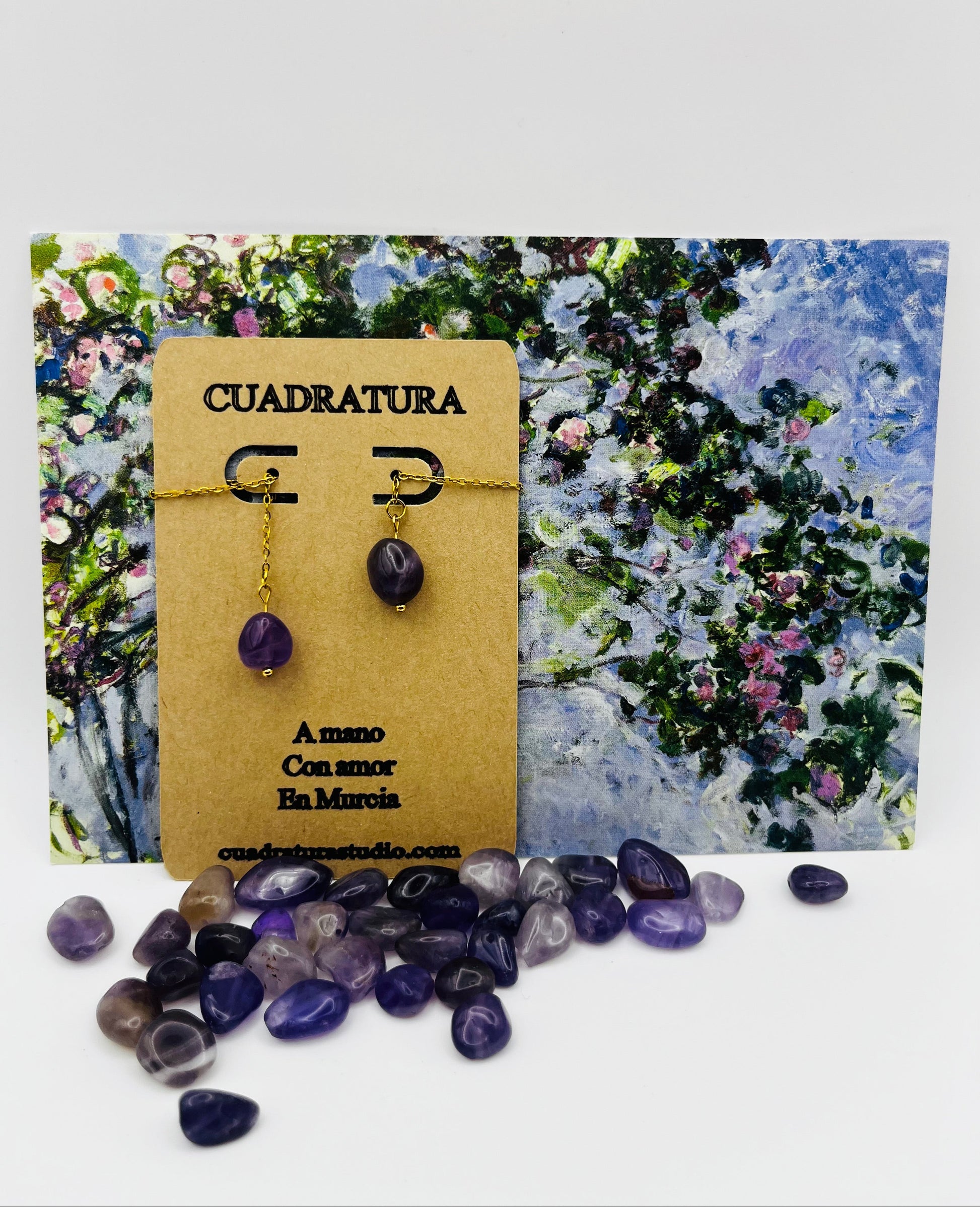 Pendientes Délica – pendientes simétricos con gancho threader dorado, cadena dorada fina y cuenta ovalada de amatista morada natural, en tarjeta Cuadratura, sobre postal de pintura impresionista de jardín con flores lilas y azules, con cuentas de amatista morada sueltas en la base, hechos a mano en Murcia