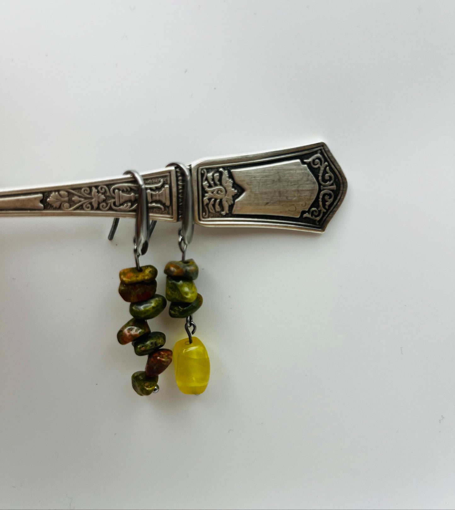 Pendientes Elena – dos aros plateados con chips de unakita verde oliva y marrón apilados y cuenta ovalada de ámbar amarillo natural colgando, colgados del mango de un cubierto de plata antigua labrada con motivos florales, fondo blanco, hechos a mano en Murcia