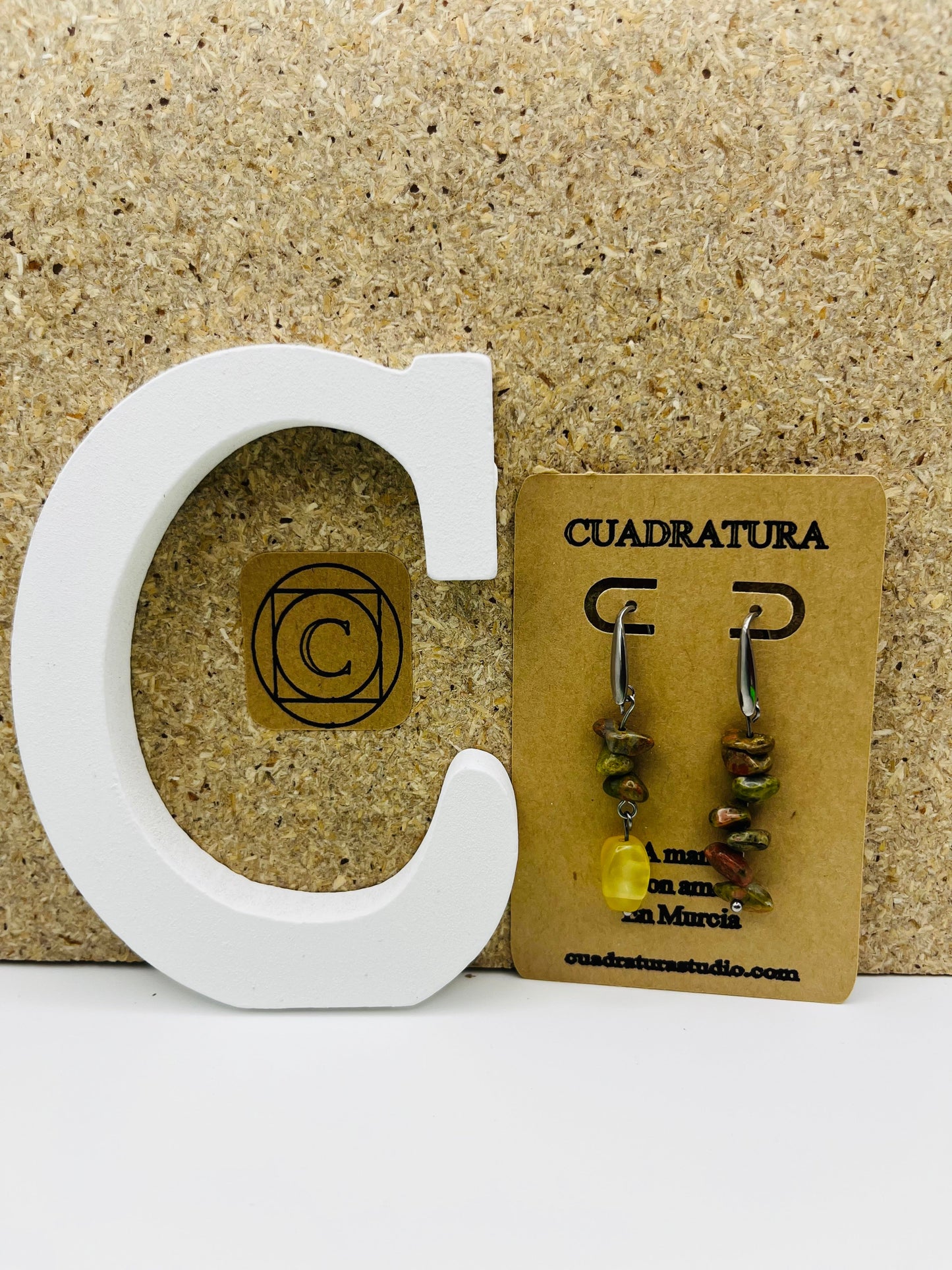 Pendientes Elena – pendientes simétricos con aro plateado, chips de unakita verde oliva y marrón apilados y cuenta ovalada de ámbar amarillo natural colgando, en tarjeta Cuadratura apoyada en letra C blanca, fondo de corcho, hechos a mano en Murcia