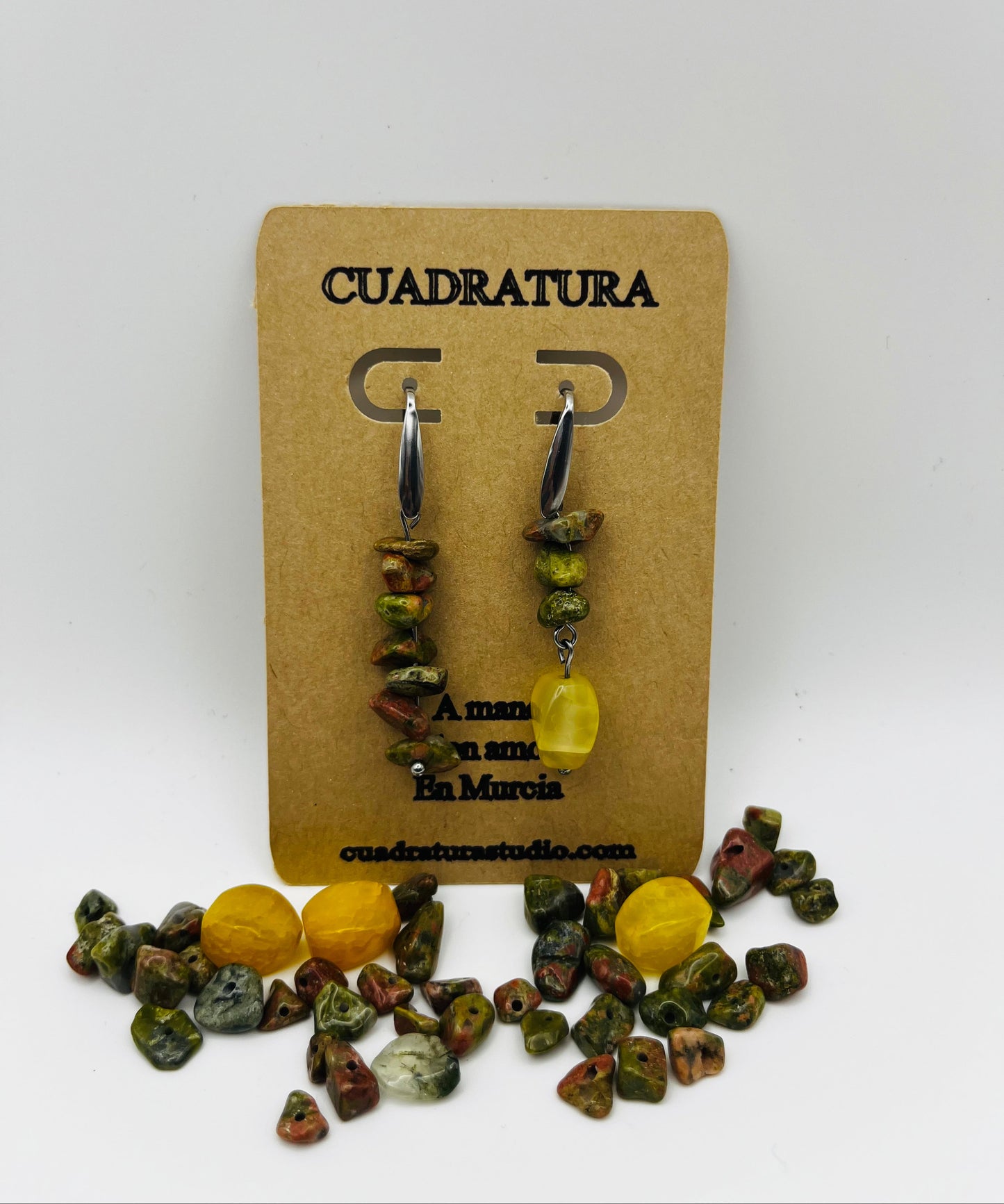 Pendientes Elena – pendientes simétricos con aro plateado, chips de unakita verde oliva y marrón apilados y cuenta ovalada de ámbar amarillo natural colgando, en tarjeta Cuadratura, sobre montículo de chips de unakita y cuentas de ámbar amarillo sueltas, fondo blanco, hechos a mano en Murcia