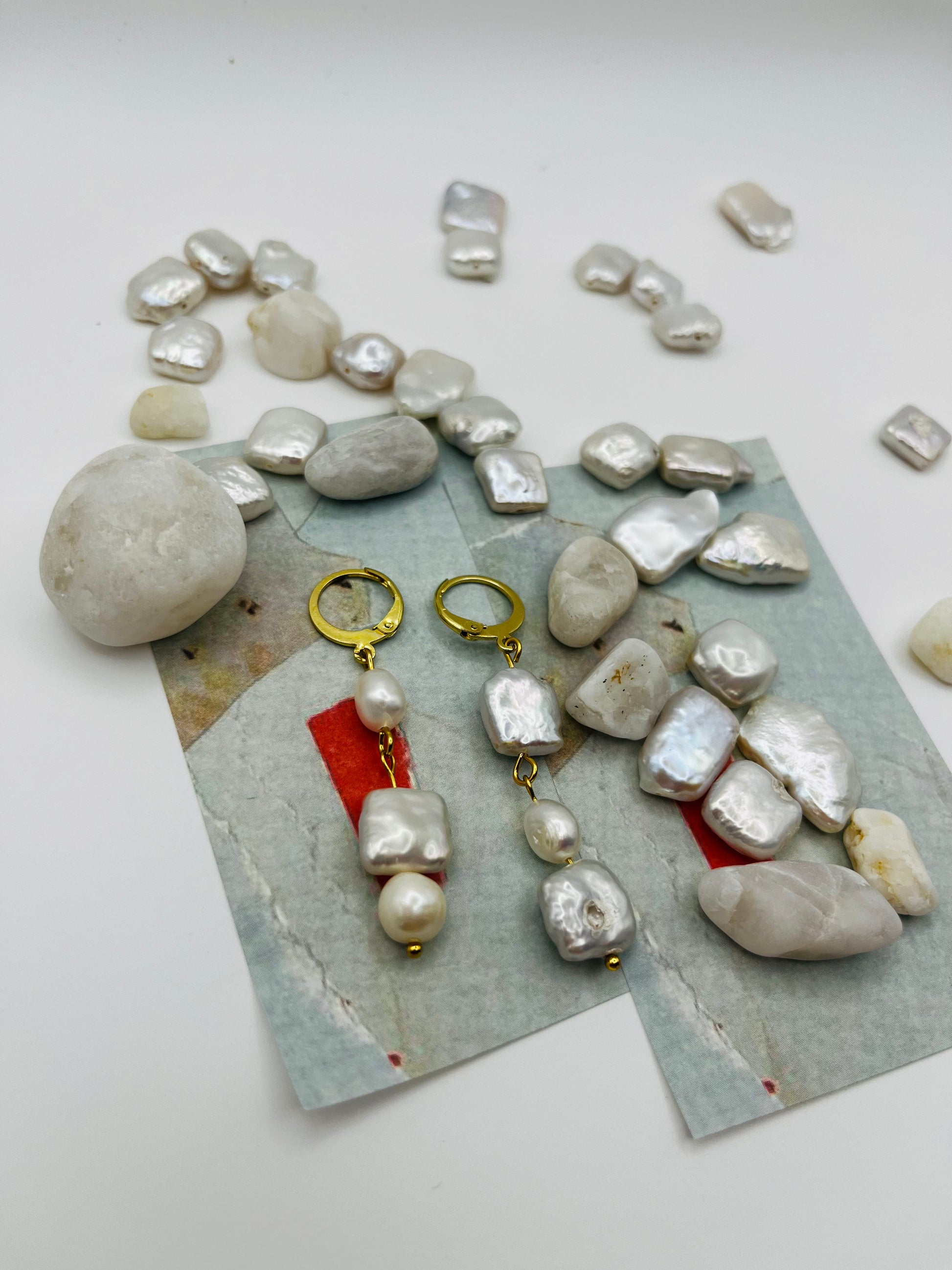 Pendientes Espuma – pendientes con aro dorado y tres perlas barrocas de agua dulce blancas en cadena (keshi, ovalada y cuadrada), sobre papel de acuarela azul grisáceo con perlas barrocas cuadradas sueltas y piedras de playa blancas esparcidas, hechos a mano en Murcia