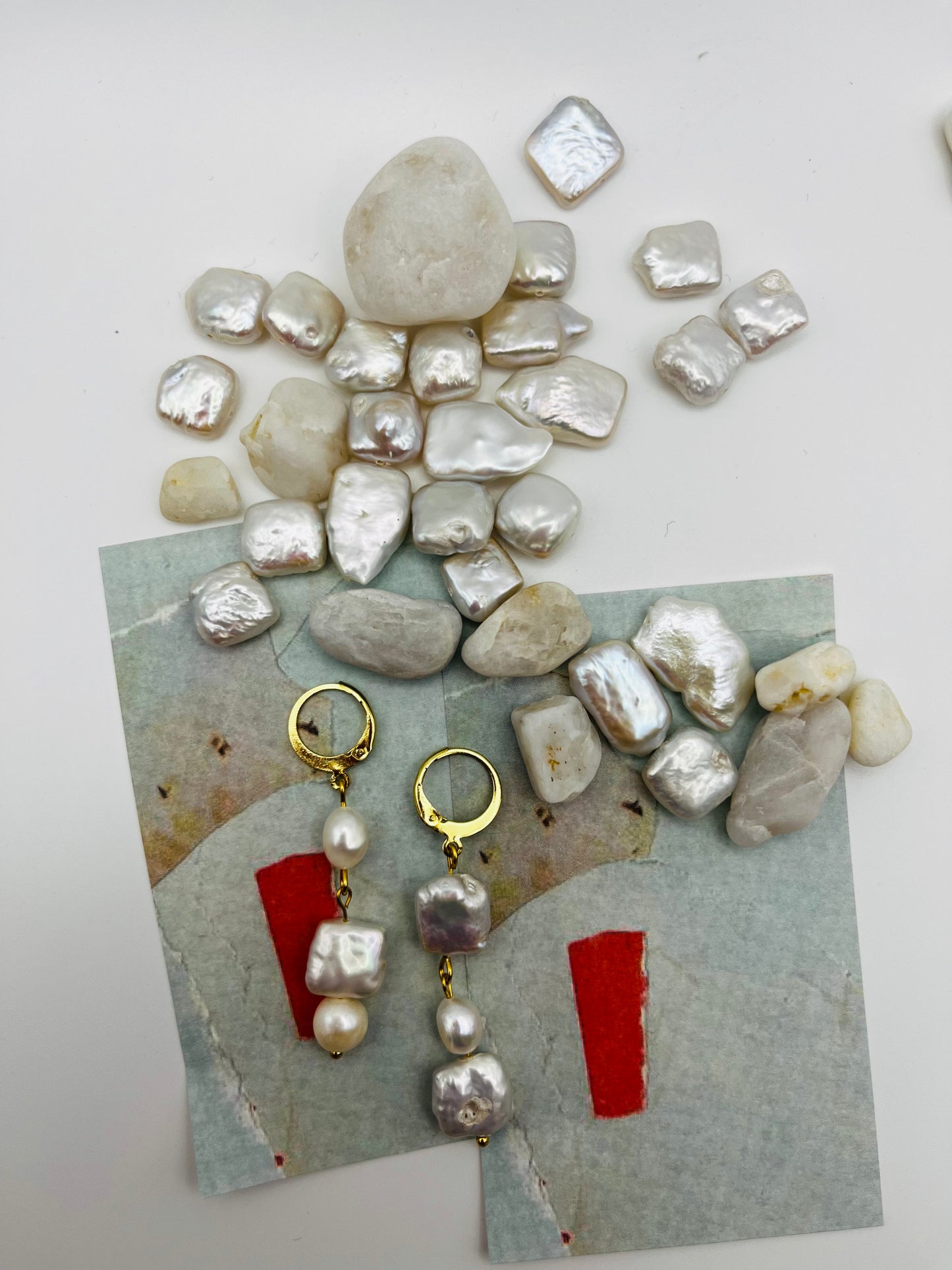 Pendientes Espuma – pendientes con aro dorado y tres perlas barrocas de agua dulce blancas en cadena (keshi, ovalada y cuadrada), sobre papel de acuarela azul grisáceo con perlas barrocas cuadradas sueltas y piedras de playa blancas esparcidas, hechos a mano en Murcia