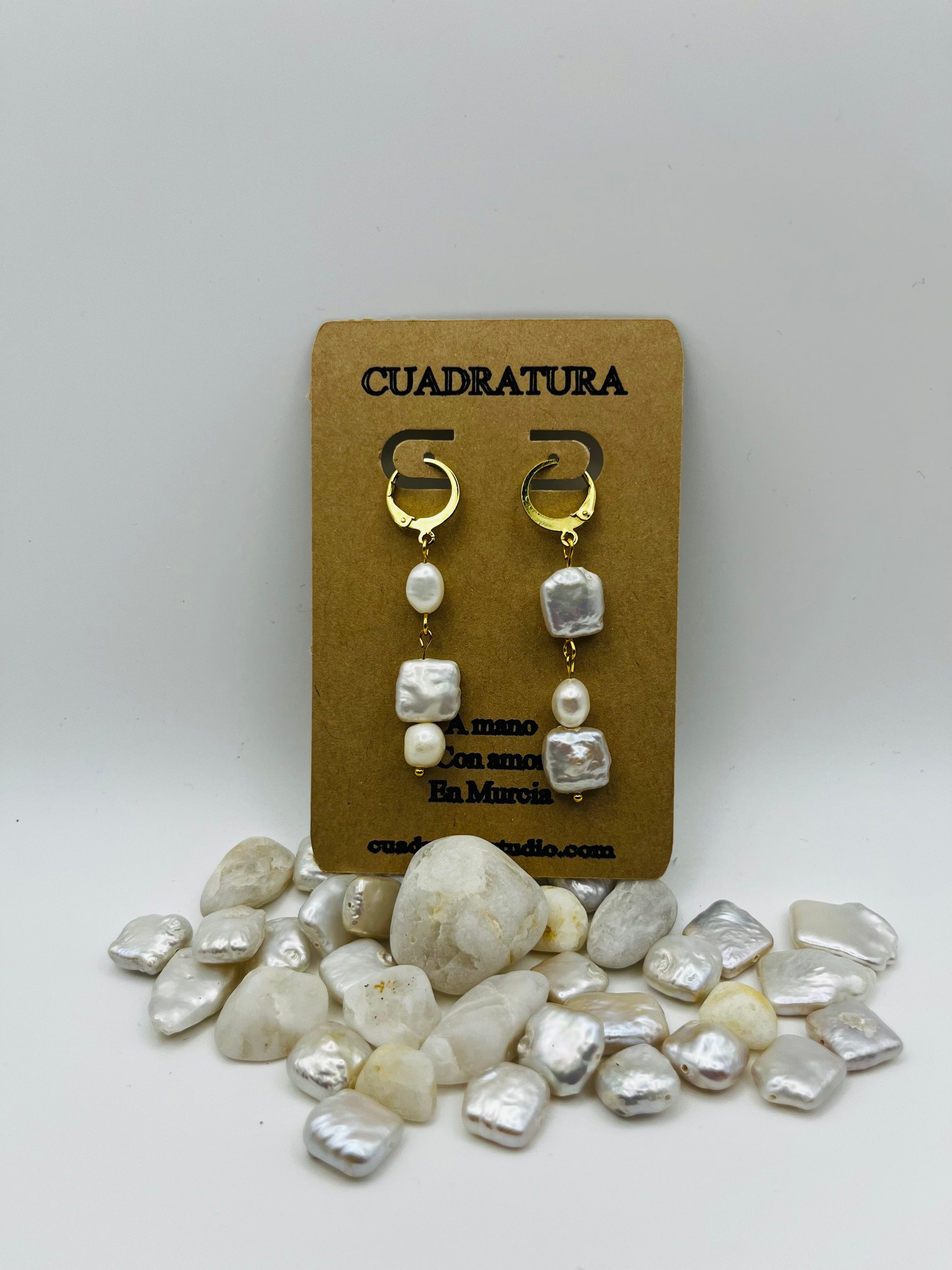 Pendientes Espuma – pendientes con aro dorado y tres perlas barrocas de agua dulce blancas en cadena (keshi, ovalada y cuadrada), en tarjeta Cuadratura, sobre montículo de perlas barrocas cuadradas y piedras blancas, fondo gris claro, hechos a mano en Murcia