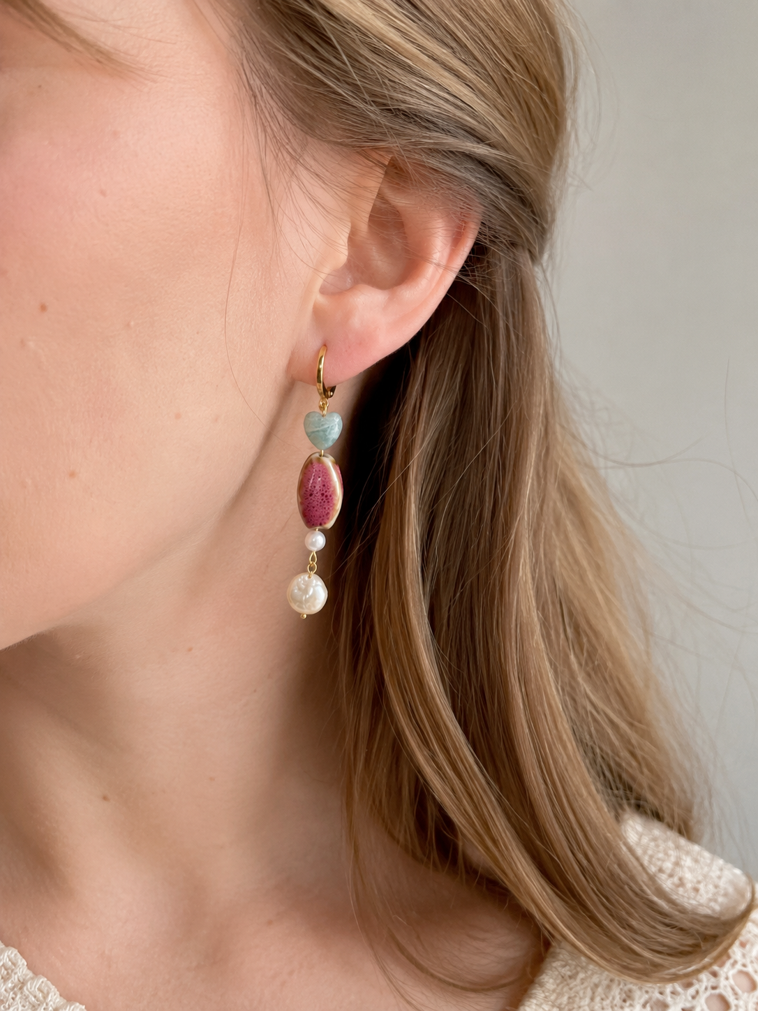 Mujer luciendo un pendiente Gaia hecho a mano con corazón de amazonita, cerámica artesanal rosa, perlas de agua dulce y aro de acero inoxidable dorado.