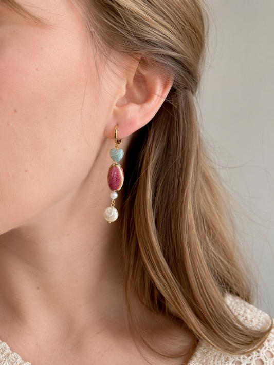 Mujer luciendo un pendiente Gaia hecho a mano con corazón de amazonita, cerámica artesanal rosa, perlas de agua dulce y aro de acero inoxidable dorado.