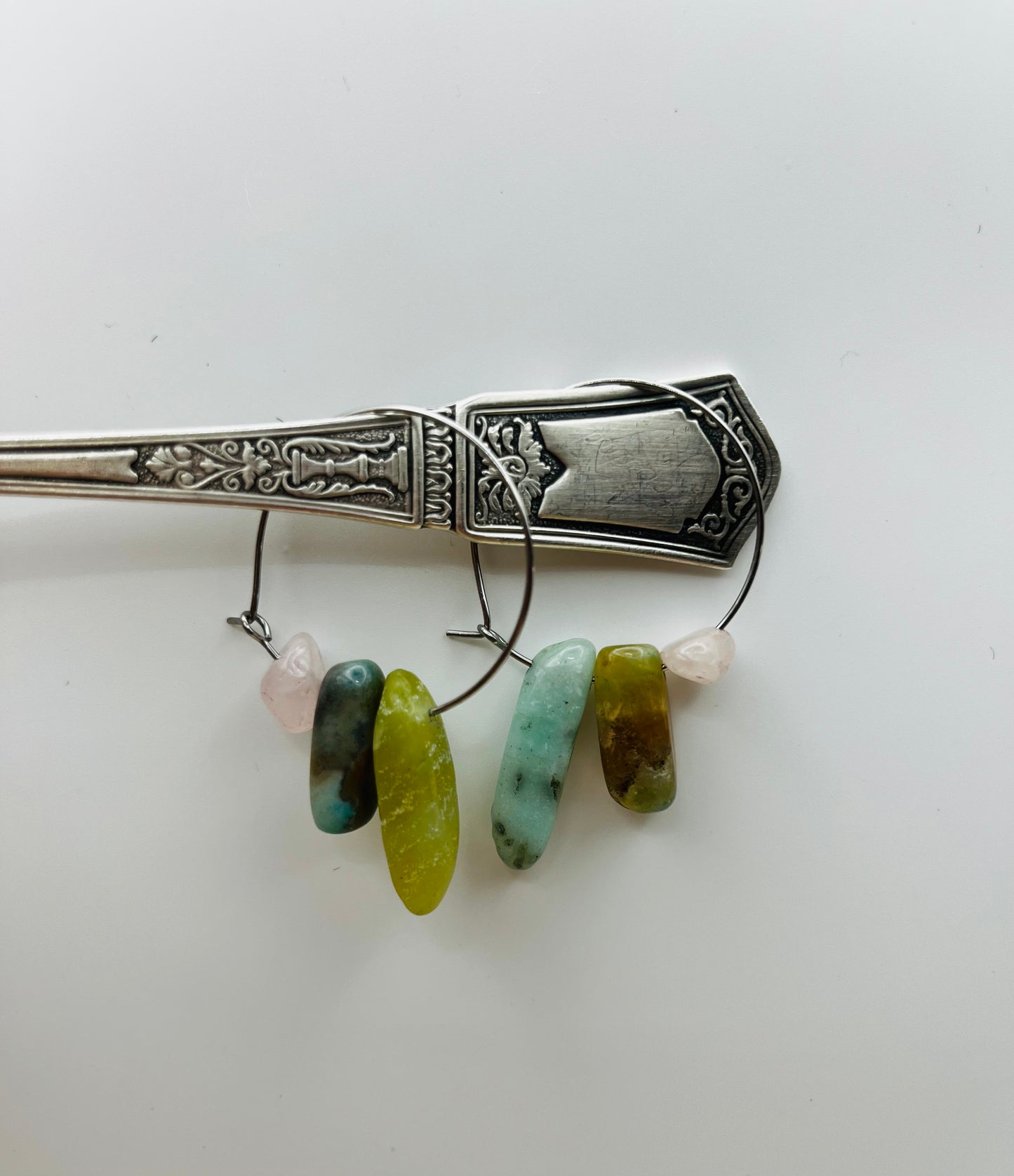 

Pendientes Levante – dos aros plateados finos con chips de jade verde oliva, jaspe marrón azulado, amazonita turquesa y cuarzo rosa lila, colgados del mango de una cuchara de plata antigua labrada, fondo blanco, hechos a mano en Murcia