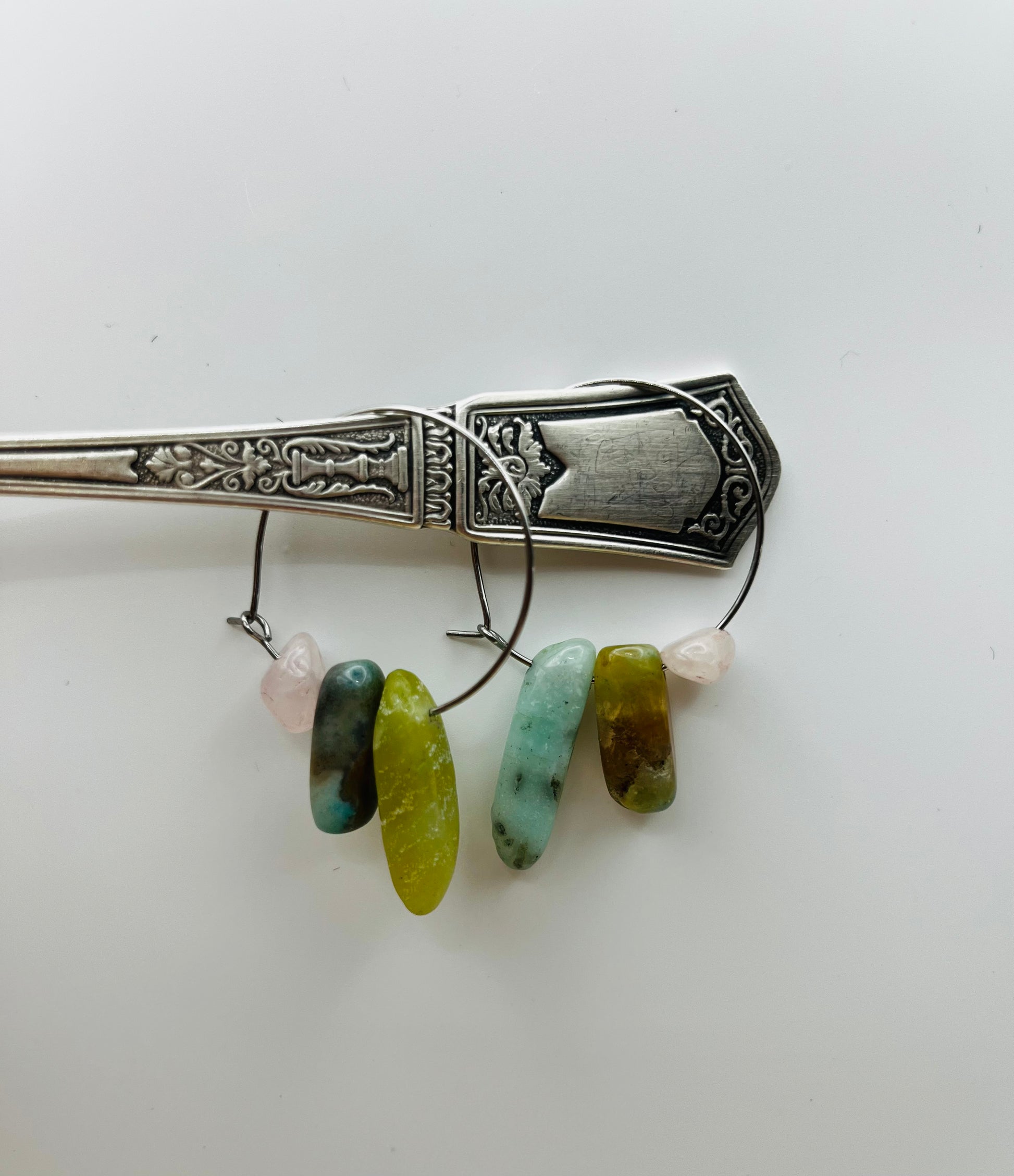

Pendientes Levante – dos aros plateados finos con chips de jade verde oliva, jaspe marrón azulado, amazonita turquesa y cuarzo rosa lila, colgados del mango de una cuchara de plata antigua labrada, fondo blanco, hechos a mano en Murcia