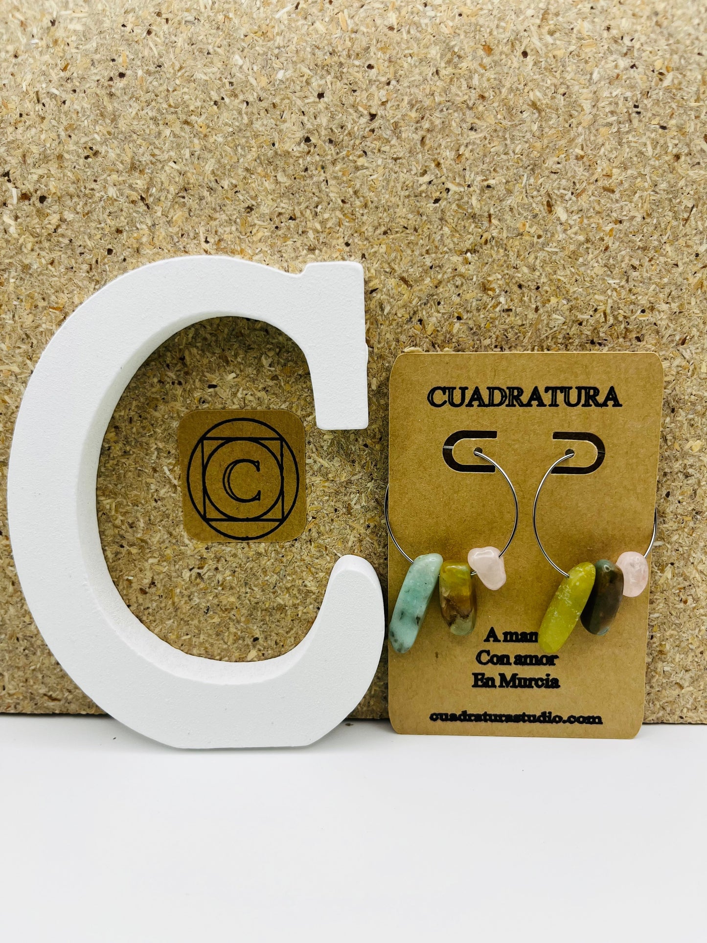 Pendientes Levante – pendientes asimétricos con aro plateado fino y chips de amazonita turquesa, jade verde oliva, cuarzo rosa y jaspe marrón, en tarjeta Cuadratura, sobre letra C blanca y fondo de corcho, hechos a mano en Murcia