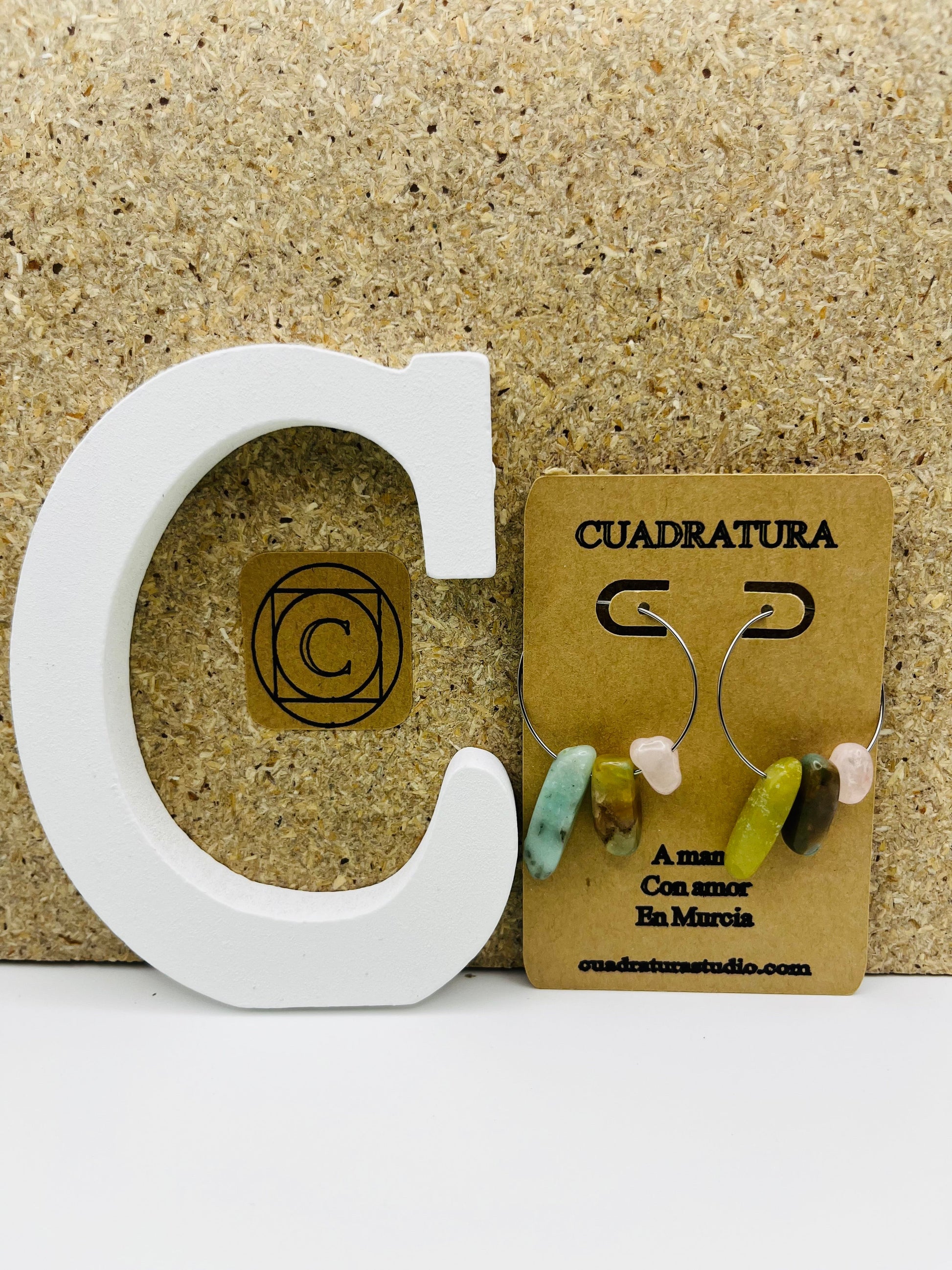 Pendientes Levante – pendientes asimétricos con aro plateado fino y chips de amazonita turquesa, jade verde oliva, cuarzo rosa y jaspe marrón, en tarjeta Cuadratura, sobre letra C blanca y fondo de corcho, hechos a mano en Murcia