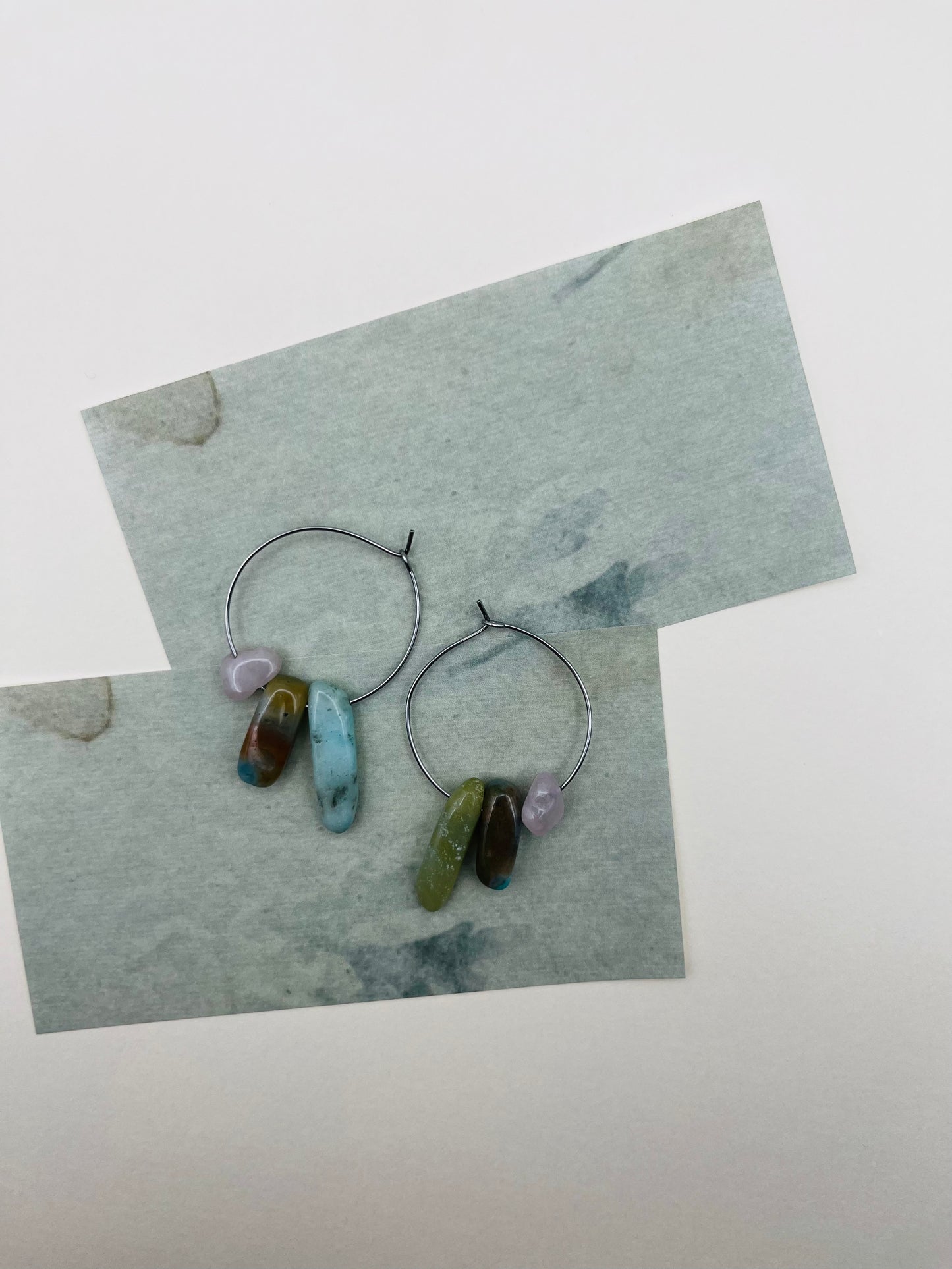 Pendientes Levante – dos aros plateados finos con chips de amazonita turquesa, jade verde oliva, jaspe marrón y cuarzo rosa, desplegados sobre tarjetas de papel marmolado azul grisáceo superpuestas, fondo blanco, hechos a mano en Murcia
