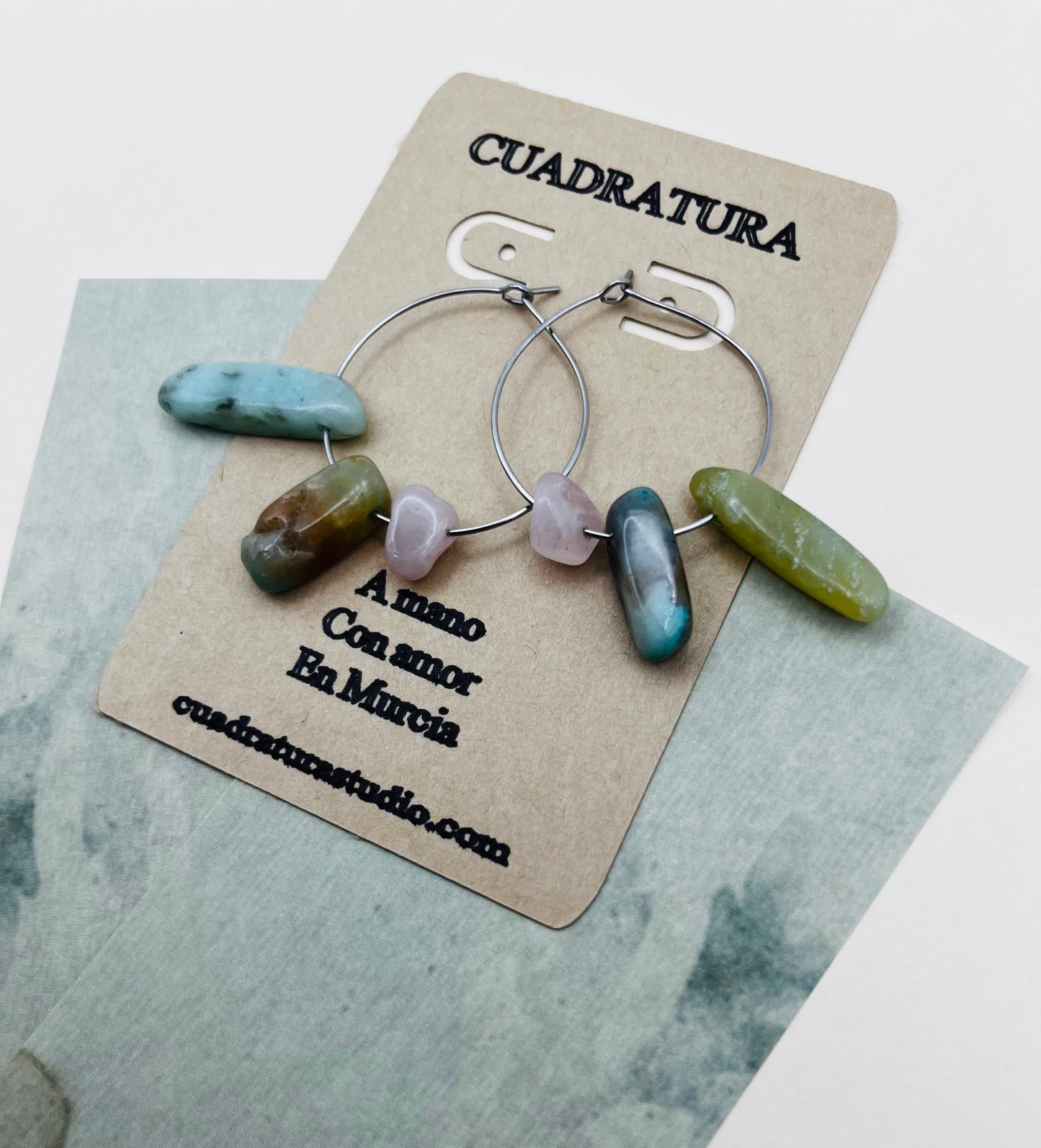 Pendientes Levante – dos aros plateados finos con chips de amazonita turquesa, jaspe marrón azulado, cuarzo rosa lila y jade verde oliva, en tarjeta Cuadratura, sobre papel marmolado azul grisáceo, hechos a mano en Murcia