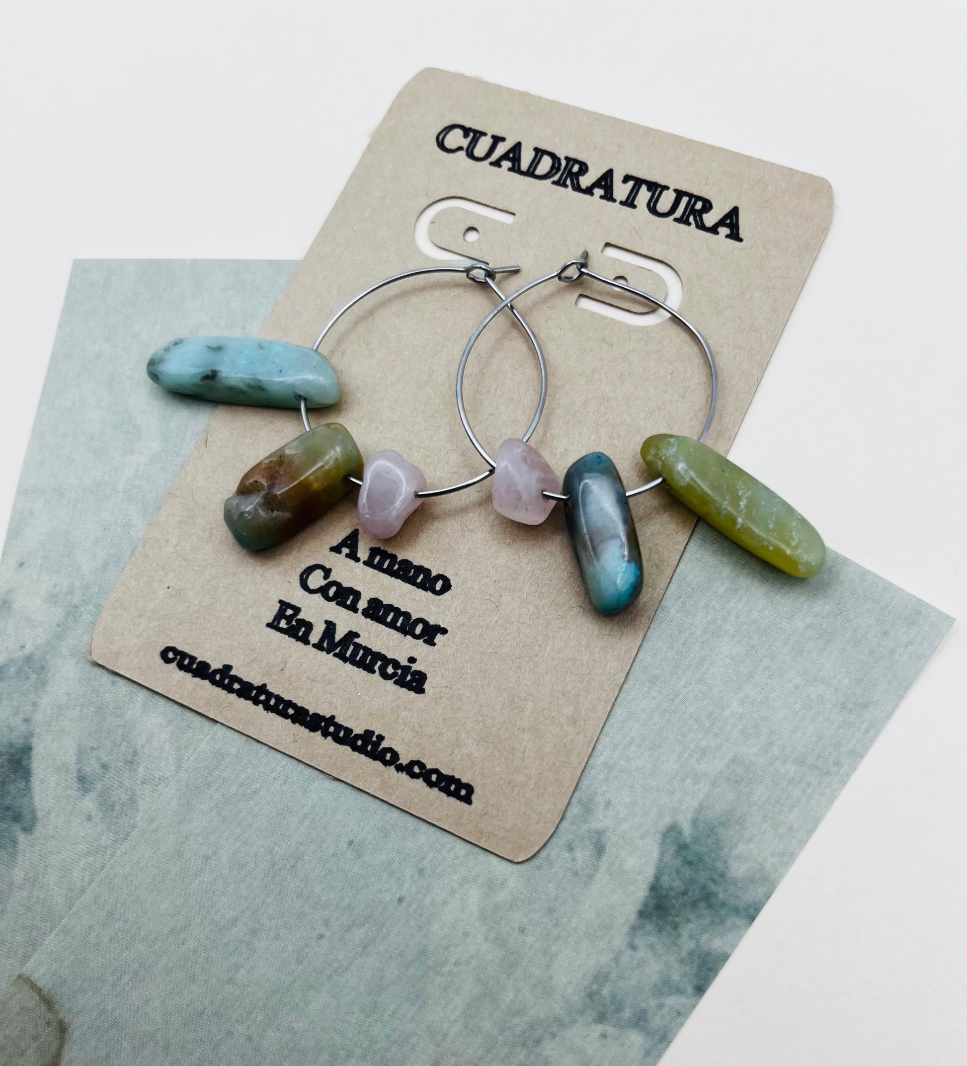 Pendientes Levante – dos aros plateados finos con chips de amazonita turquesa, jaspe marrón azulado, cuarzo rosa lila y jade verde oliva, en tarjeta Cuadratura, sobre papel marmolado azul grisáceo, hechos a mano en Murcia