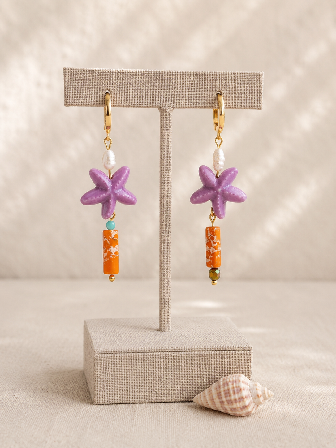 Pendientes artesanales Mediterráneo sobre expositor de joyería. Aros de acero inoxidable chapados en oro con perlas de agua dulce, estrellas de mar de cerámica esmaltada lila y barras de jaspe sedimentario naranja, en diseño asimétrico junto a una caracola.