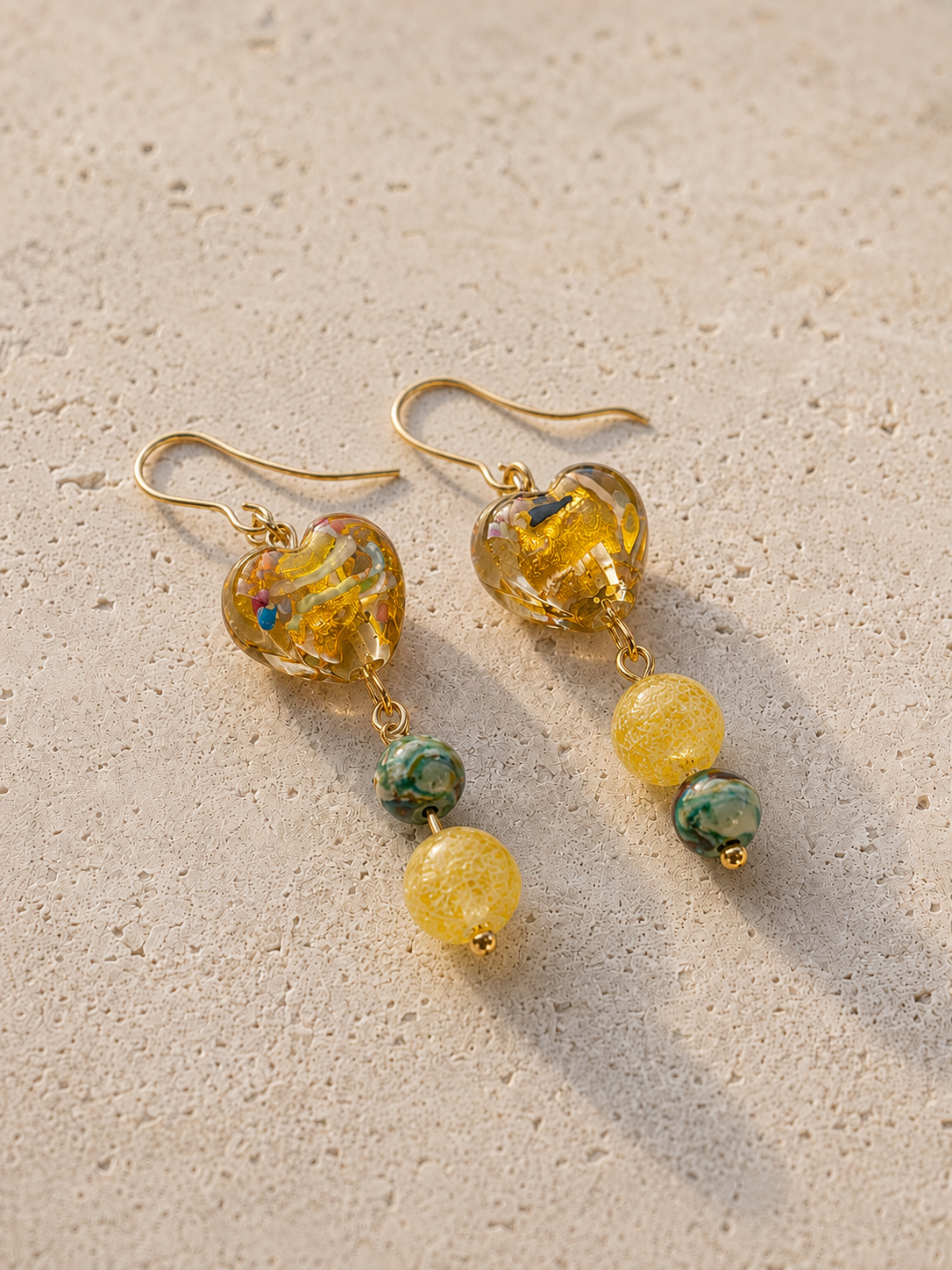 Pendientes Miel hechos a mano con corazones de cristal dorado, cuentas amarillas y piedras verdes, sobre fondo de piedra beige.
