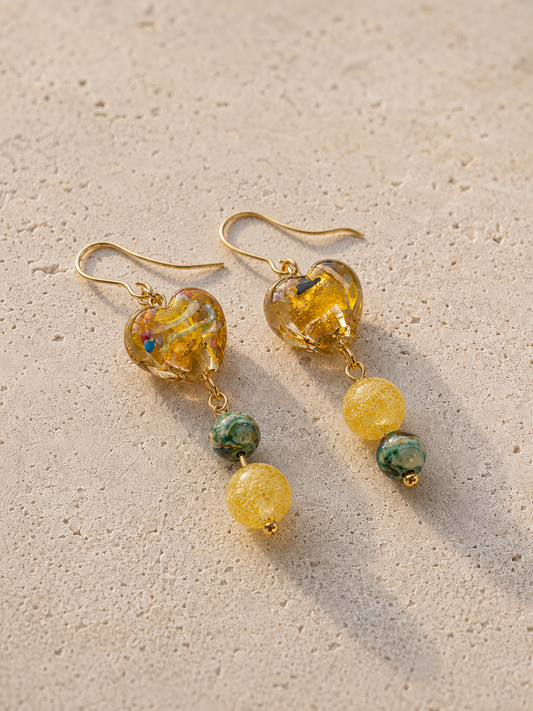 Pendientes Miel hechos a mano con corazones de cristal dorado, cuentas amarillas y piedras verdes, sobre fondo de piedra beige.