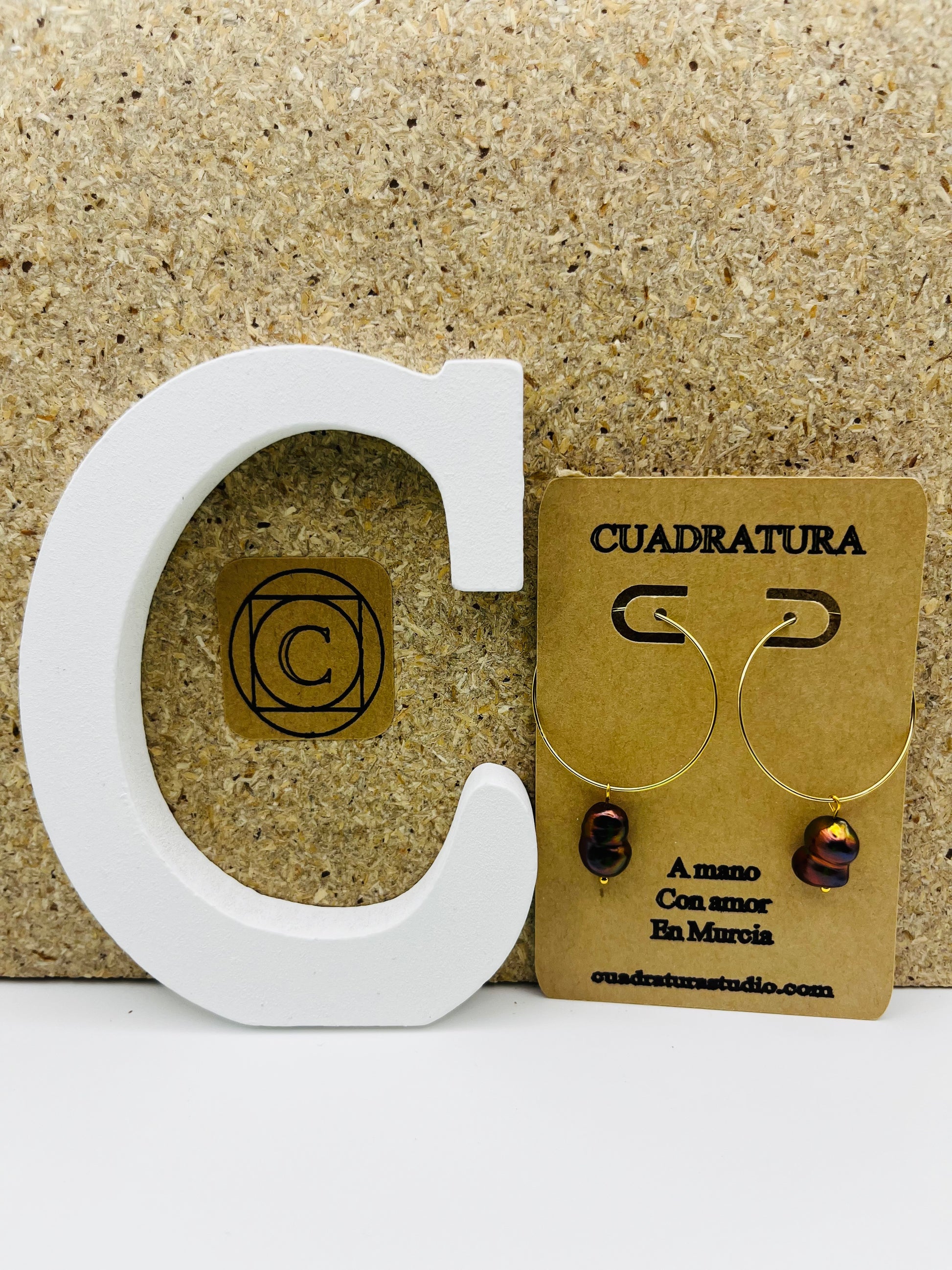 Pendientes Peacock – pendientes simétricos con aro dorado fino grande y perla keshi marrón oscuro iridiscente colgando, en tarjeta Cuadratura apoyada en letra C blanca, fondo de corcho, hechos a mano en Murcia