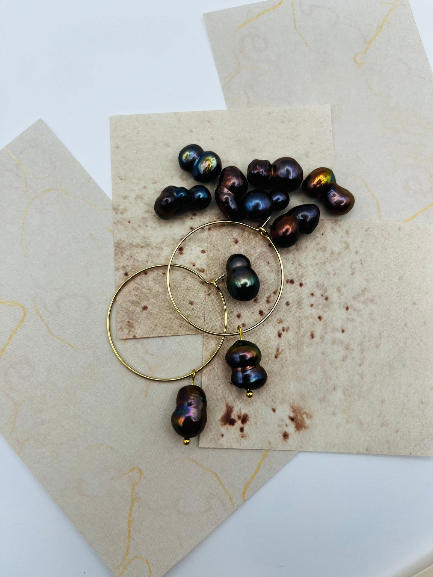 Pendientes Peacock – dos aros dorados finos grandes con perla keshi marrón oscuro iridiscente tornasolada en azul, verde y bronce, desplegados sobre hojas de papel marmolado crema superpuestas, con perlas keshi sueltas esparcidas, fondo blanco, hechos a mano en Murcia