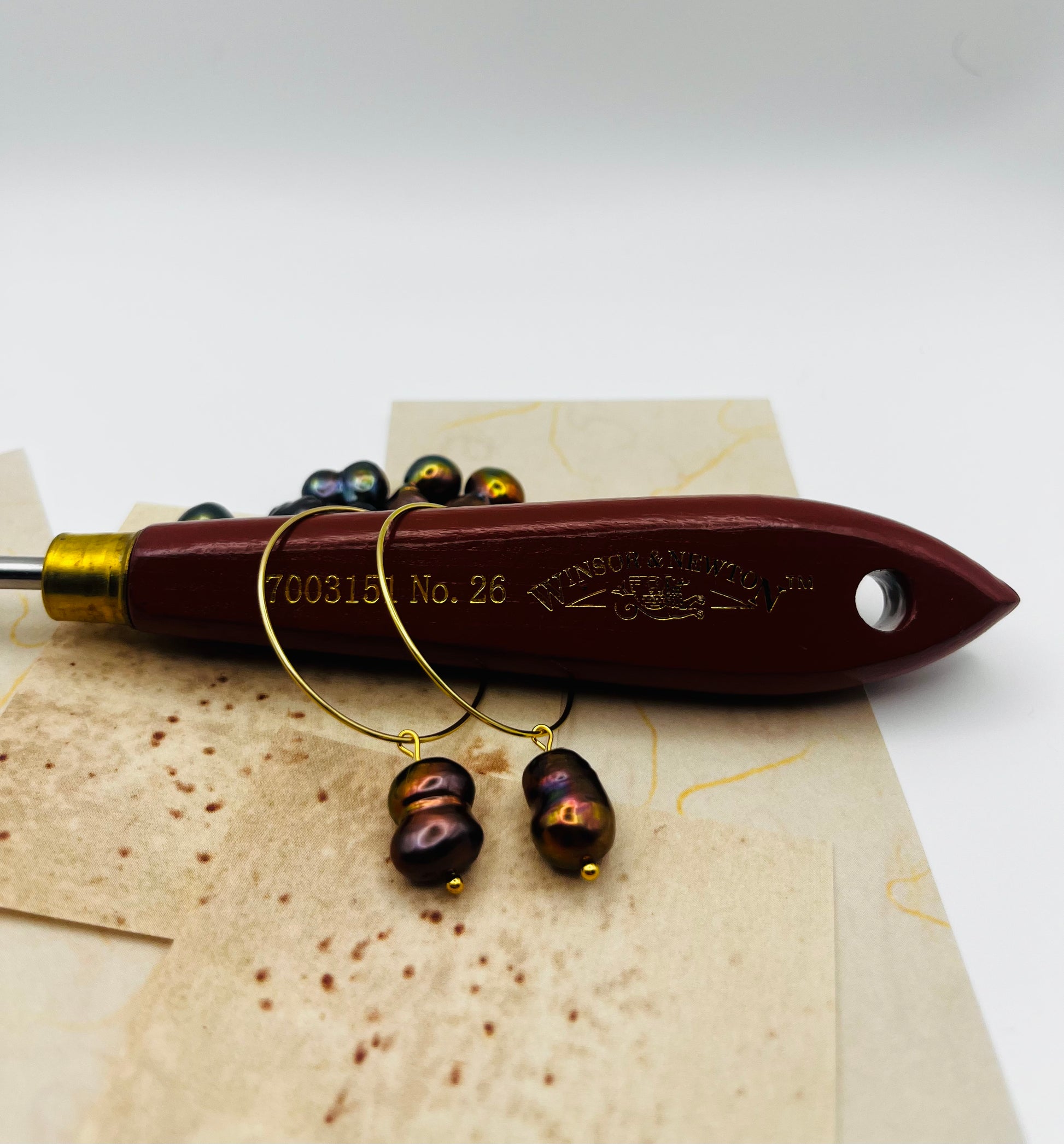 Pendientes Peacock – dos aros dorados finos grandes con perla keshi marrón oscuro iridiscente tornasolada en azul, verde y bronce, colgados de pincel Winsor & Newton No. 26 con mango granate, sobre hojas de papel marmolado crema, con perlas keshi sueltas encima del pincel, fondo blanco, hechos a mano en Murcia