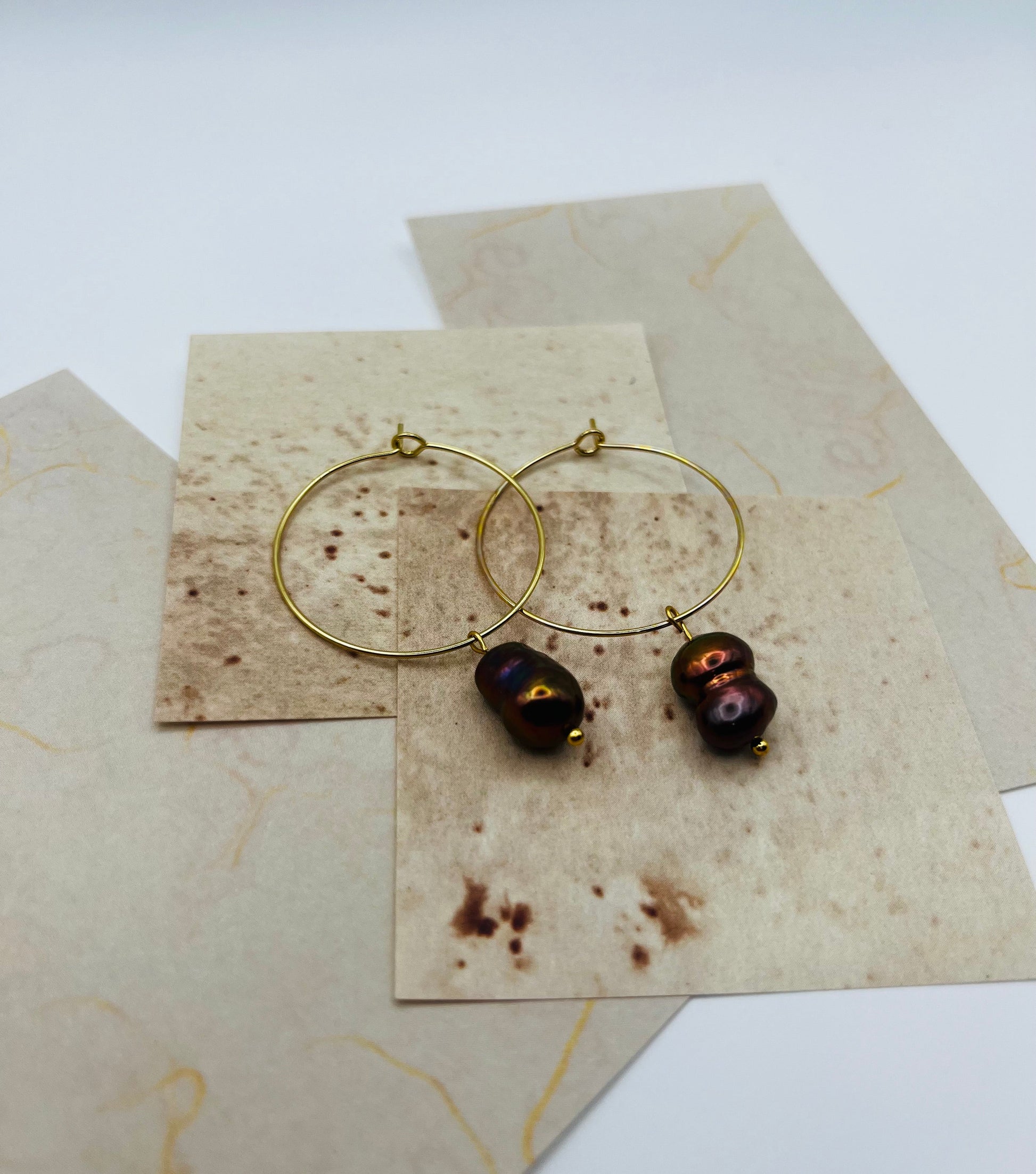 Pendientes Peacock – dos aros dorados finos grandes con perla keshi marrón oscuro iridiscente tornasolada en azul, verde y bronce, en plano cenital sobre hojas de papel marmolado crema superpuestas, fondo blanco, hechos a mano en Murcia