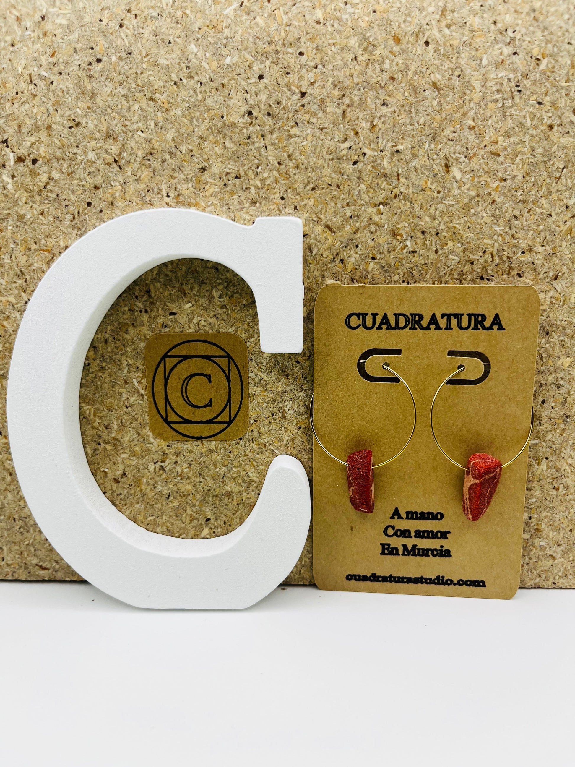 Pendientes Peñón – pendientes simétricos con aro dorado fino y pieza de coral rojo natural, en tarjeta Cuadratura, sobre letra C blanca y fondo de corcho, hechos a mano en Murcia