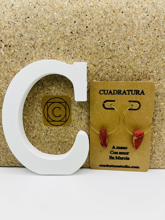 Pendientes Peñón – pendientes simétricos con aro dorado fino y pieza de coral rojo natural, en tarjeta Cuadratura, sobre letra C blanca y fondo de corcho, hechos a mano en Murcia