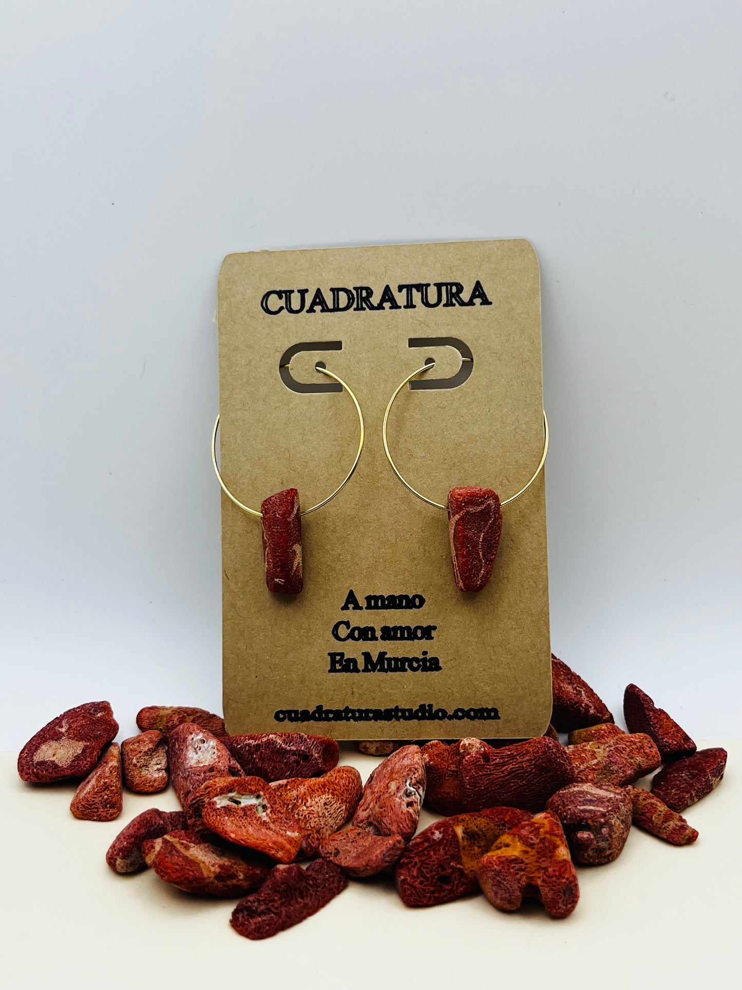 Pendientes Peñón – pendientes simétricos con aro dorado fino y pieza de coral rojo natural rugoso, en tarjeta Cuadratura, sobre montículo de trozos de coral rojo natural de distintas formas, fondo gris claro, hechos a mano en Murcia