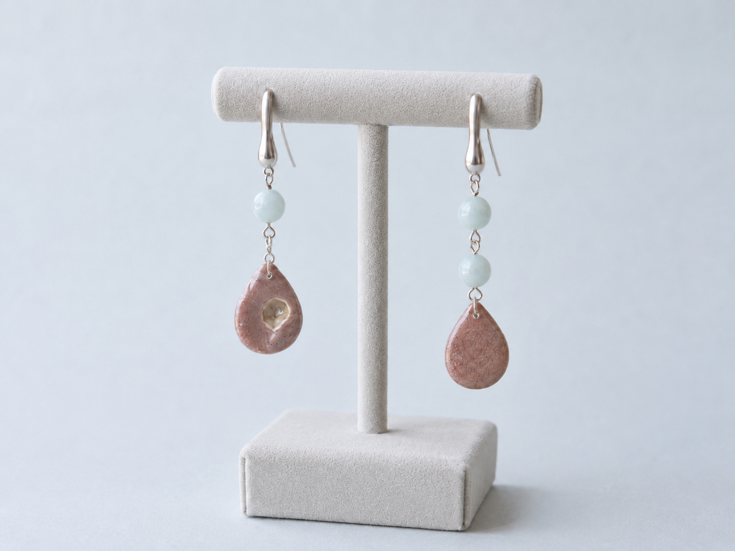 Par de pendientes artesanales Periwinkle sobre expositor de joyería, con perlas nacaradas iridiscentes en forma de gota y cuentas de piedra azul, montados en acero inoxidable y mostrando el diseño asimétrico de ambas piezas.