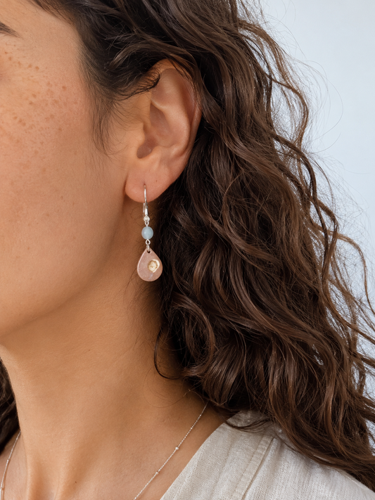 endientes artesanales Periwinkle con perla nacarada iridiscente en forma de gota y cuenta de piedra azul, montados en acero inoxidable, sobre modelo de pelo castaño rizado.