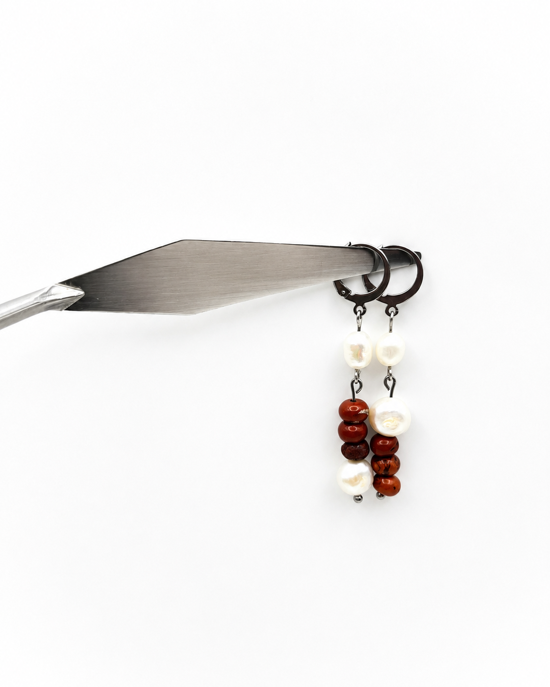 Pendientes asimétricos de Cuadratura Studio colgando de una espátula de acero: aro plateado con perla barroca blanca y cuentas de jaspe rojo, y aro con perla barroca y jaspe rojo alternas, sobre fondo blanco. Montados a mano en Murcia.`
