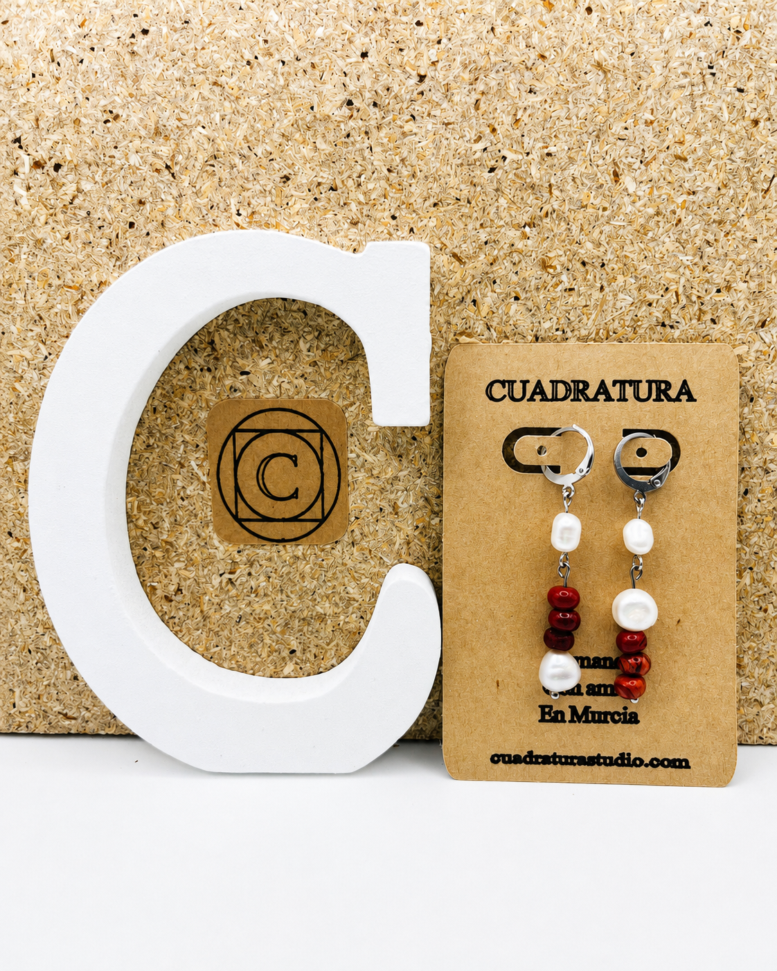 Pendientes de Cuadratura Studio en tarjeta kraft: aro de acero inoxidable plateado con perla barroca blanca, cuentas de coral rojo y segunda perla barroca, sobre fondo de corcho con letra C decorativa blanca y logo de Cuadratura. Montados a mano en Murcia.`