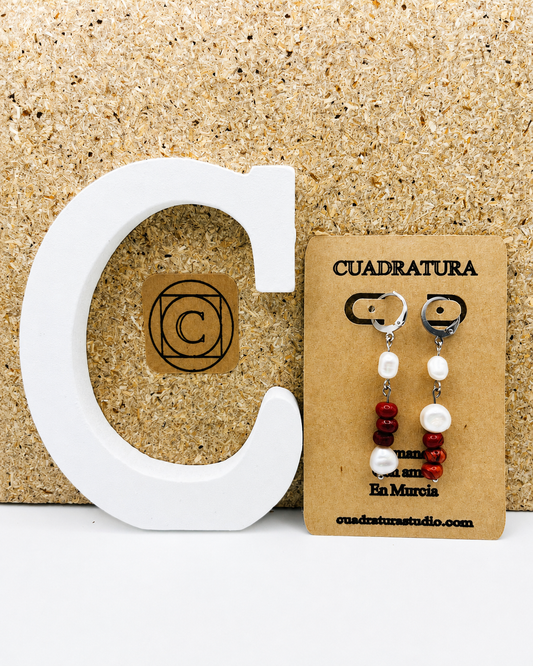Pendientes de Cuadratura Studio en tarjeta kraft: aro de acero inoxidable plateado con perla barroca blanca, cuentas de coral rojo y segunda perla barroca, sobre fondo de corcho con letra C decorativa blanca y logo de Cuadratura. Montados a mano en Murcia.`