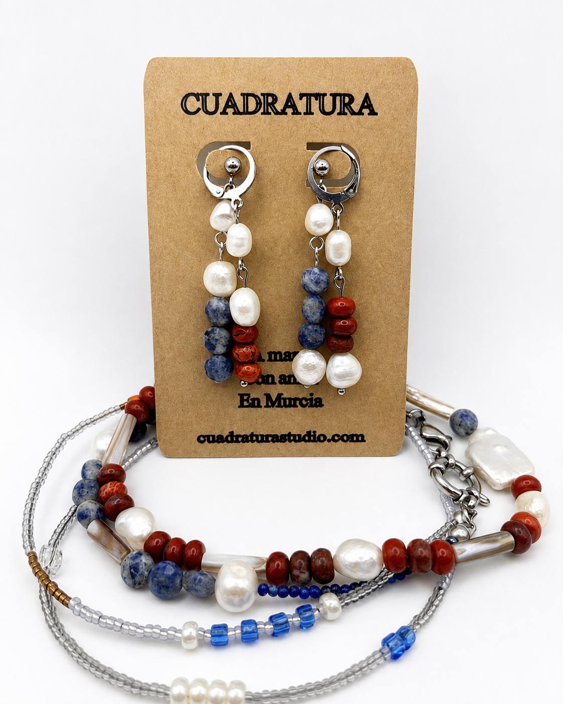 Pendientes asimétricos de Cuadratura Studio en tarjeta kraft con aro plateado, perlas barrocas blancas, cuentas de sodalita azul y jaspe rojo, junto a collar y pulsera a juego con las mismas piedras, perlas y tubos plateados. Montados a mano en Murcia.`