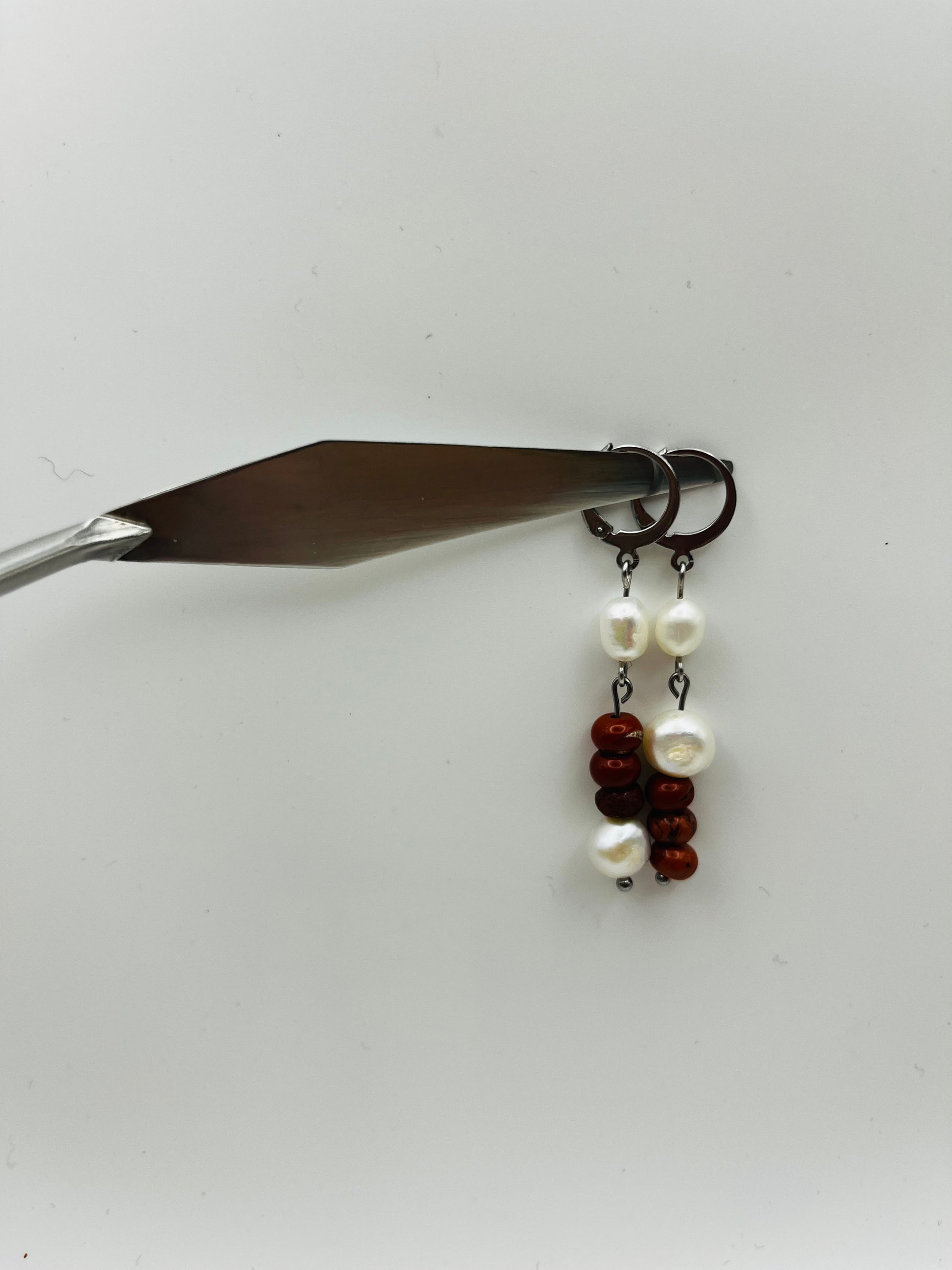 Pendientes Pleamar – dos enganches plateados tipo palanca con perla oval blanca, tres cuentas redondas de jaspe rojo oscuro y perla oval blanca en la base, colgados de espátula de pintura plateada, fondo blanco, hechos a mano en Murcia