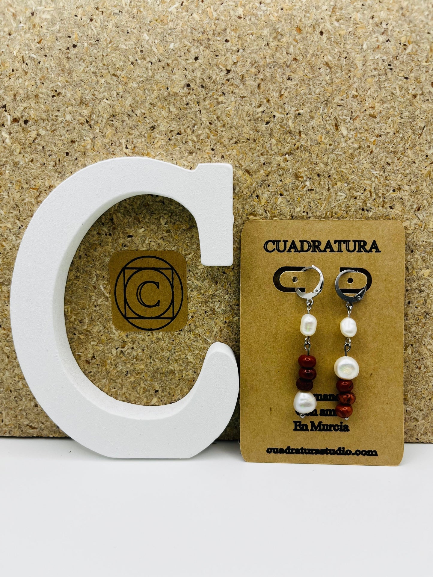 Pendientes Pleamar – pendientes simétricos con enganche plateado tipo palanca, perla oval blanca, tres cuentas redondas de jaspe rojo y perla oval blanca en la base, en tarjeta Cuadratura apoyada en letra C blanca, fondo de corcho, hechos a mano en Murcia
