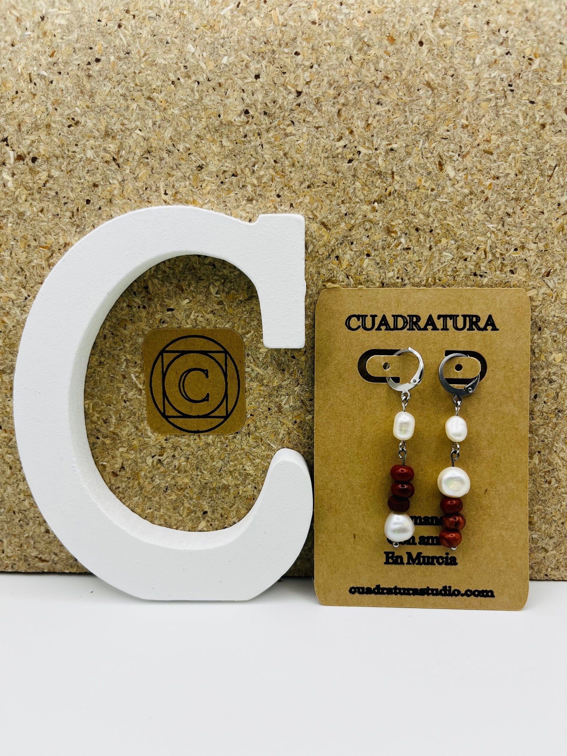 Pendientes Pleamar – pendientes simétricos con enganche plateado tipo palanca, perla oval blanca, tres cuentas redondas de jaspe rojo y perla oval blanca en la base, en tarjeta Cuadratura apoyada en letra C blanca, fondo de corcho, hechos a mano en Murcia