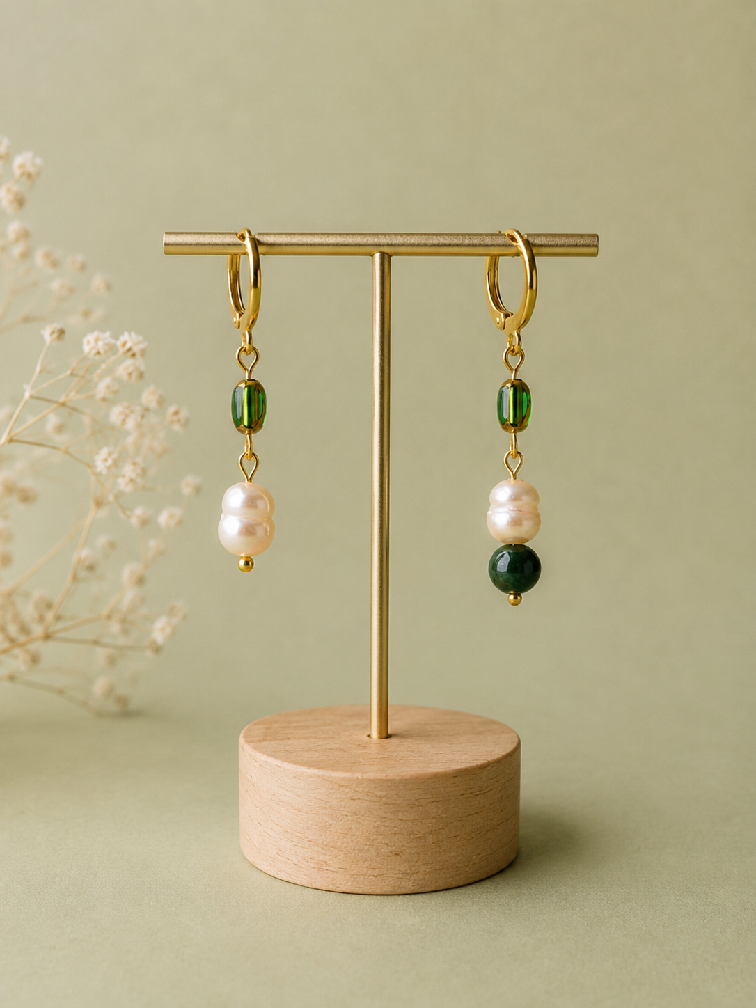 Pendientes artesanales Prao sobre expositor, con diseño asimétrico de aros de acero inoxidable dorado, cuentas de vidrio verde, perlas barrocas de agua dulce y una esfera de piedra verde en uno de los pendientes.