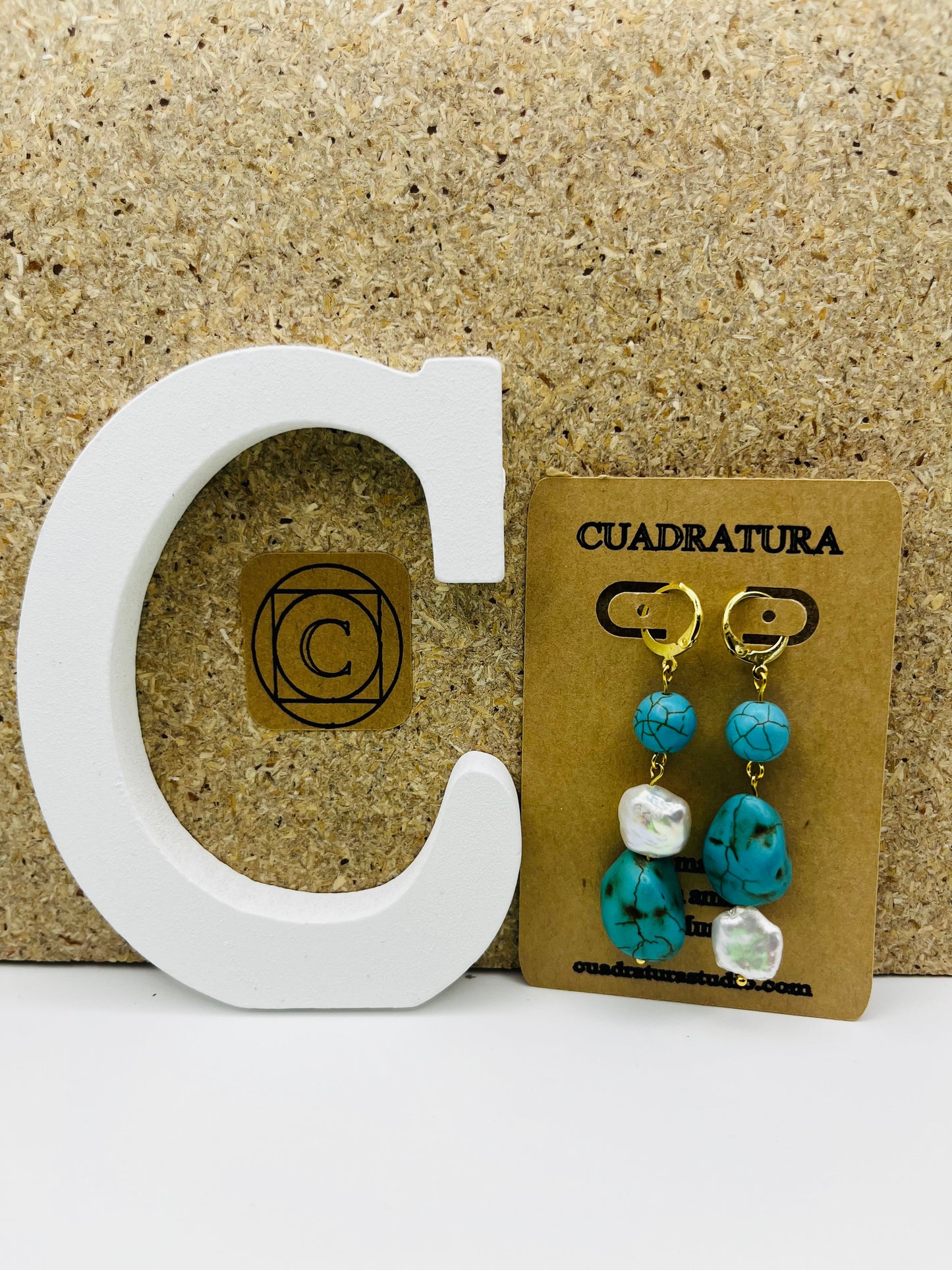 Pendientes Turquesa – pendientes simétricos con aro dorado, cuenta redonda de howlita turquesa, perla keshi blanca y cuenta grande irregular de howlita turquesa, en tarjeta Cuadratura, sobre letra C blanca y fondo de corcho, hechos a mano en Murcia