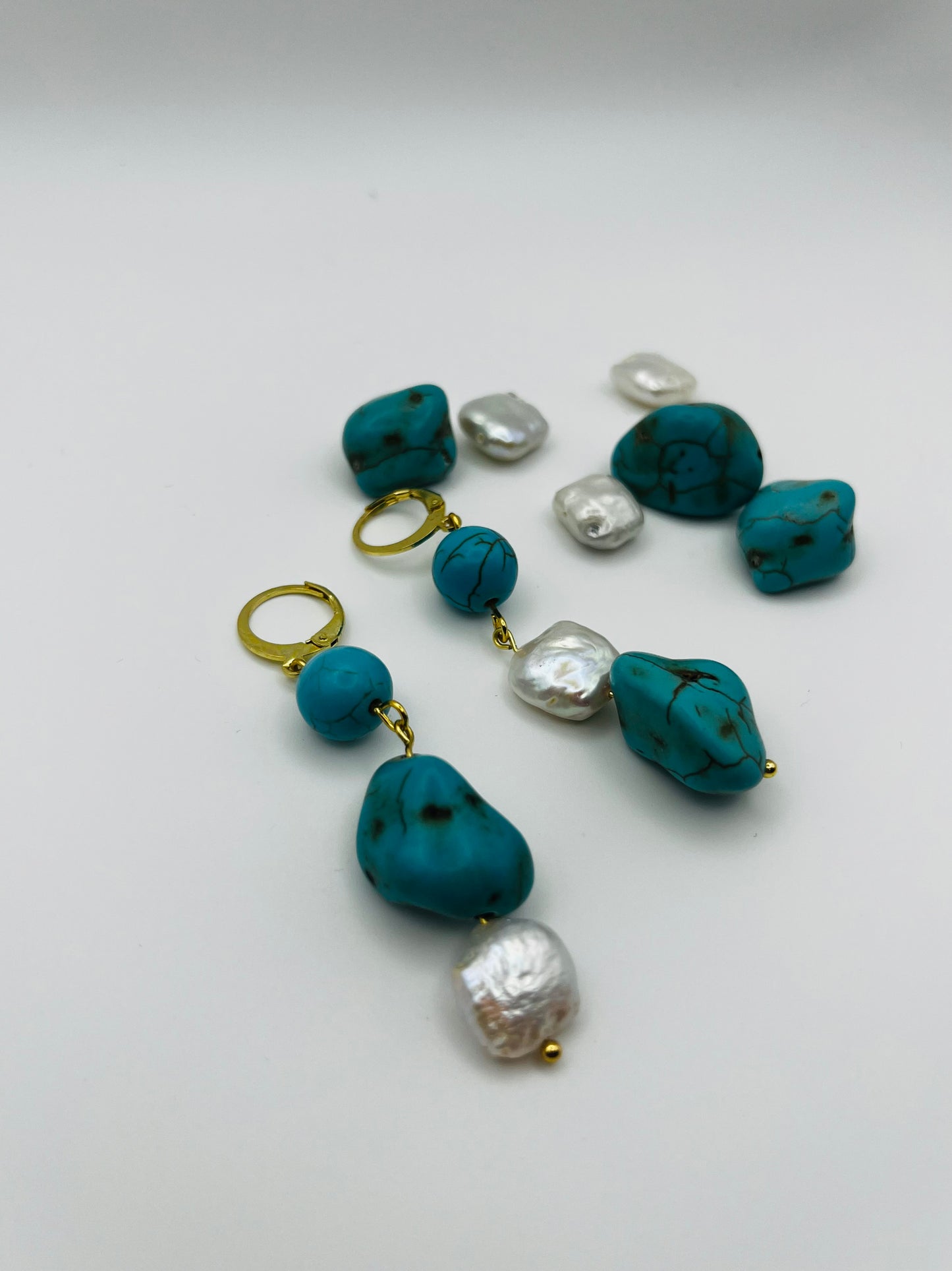 Pendientes Turquesa – pendientes simétricos con aro dorado, cuenta redonda de howlita turquesa, perla keshi blanca cuadrada y cuenta grande irregular de howlita turquesa con perla keshi blanca en la base, con cuentas de howlita turquesa y perlas keshi sueltas, sobre fondo blanco, hechos a mano en Murcia