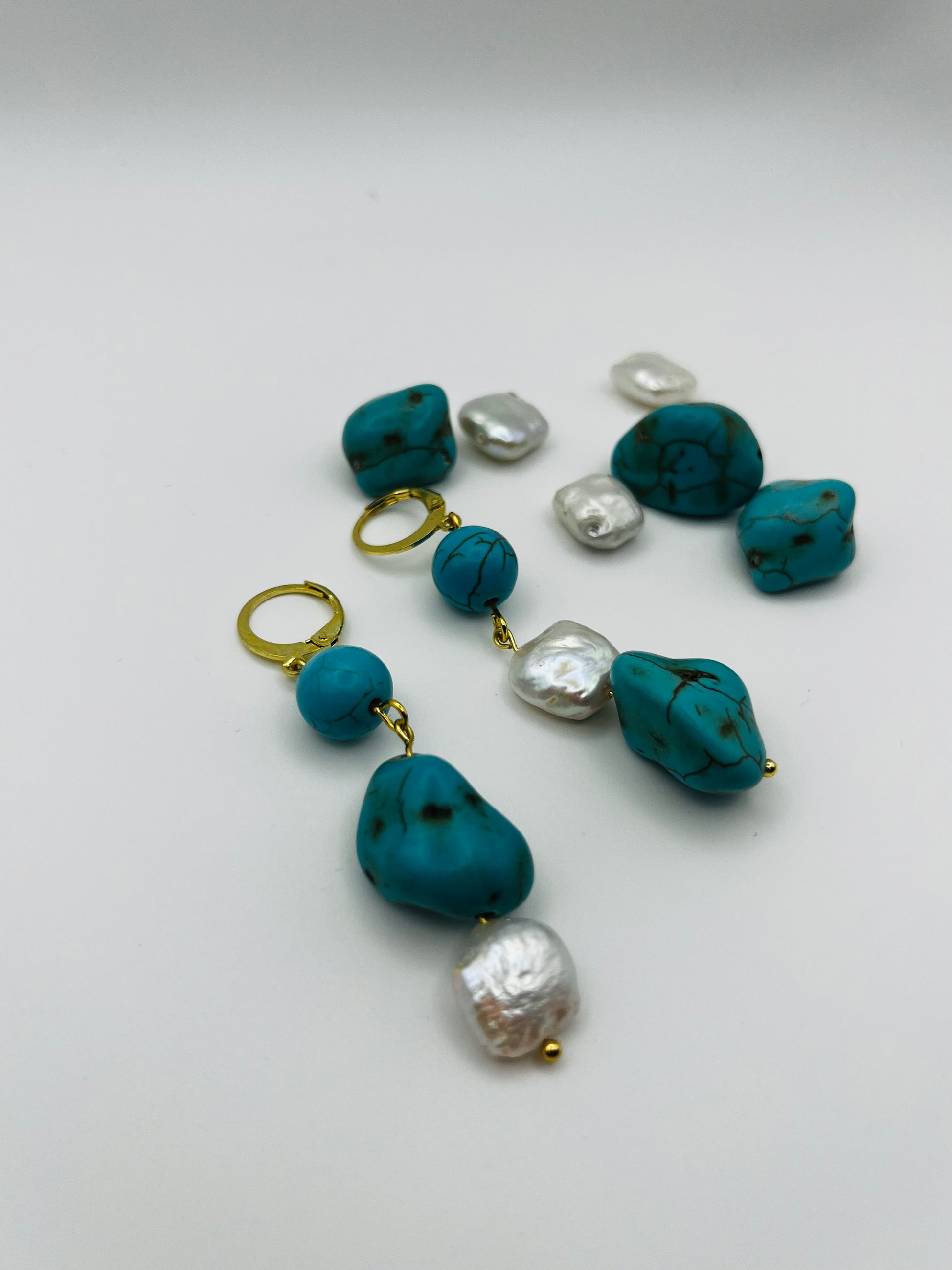 Pendientes Turquesa – pendientes simétricos con aro dorado, cuenta redonda de howlita turquesa, perla keshi blanca cuadrada y cuenta grande irregular de howlita turquesa con perla keshi blanca en la base, con cuentas de howlita turquesa y perlas keshi sueltas, sobre fondo blanco, hechos a mano en Murcia