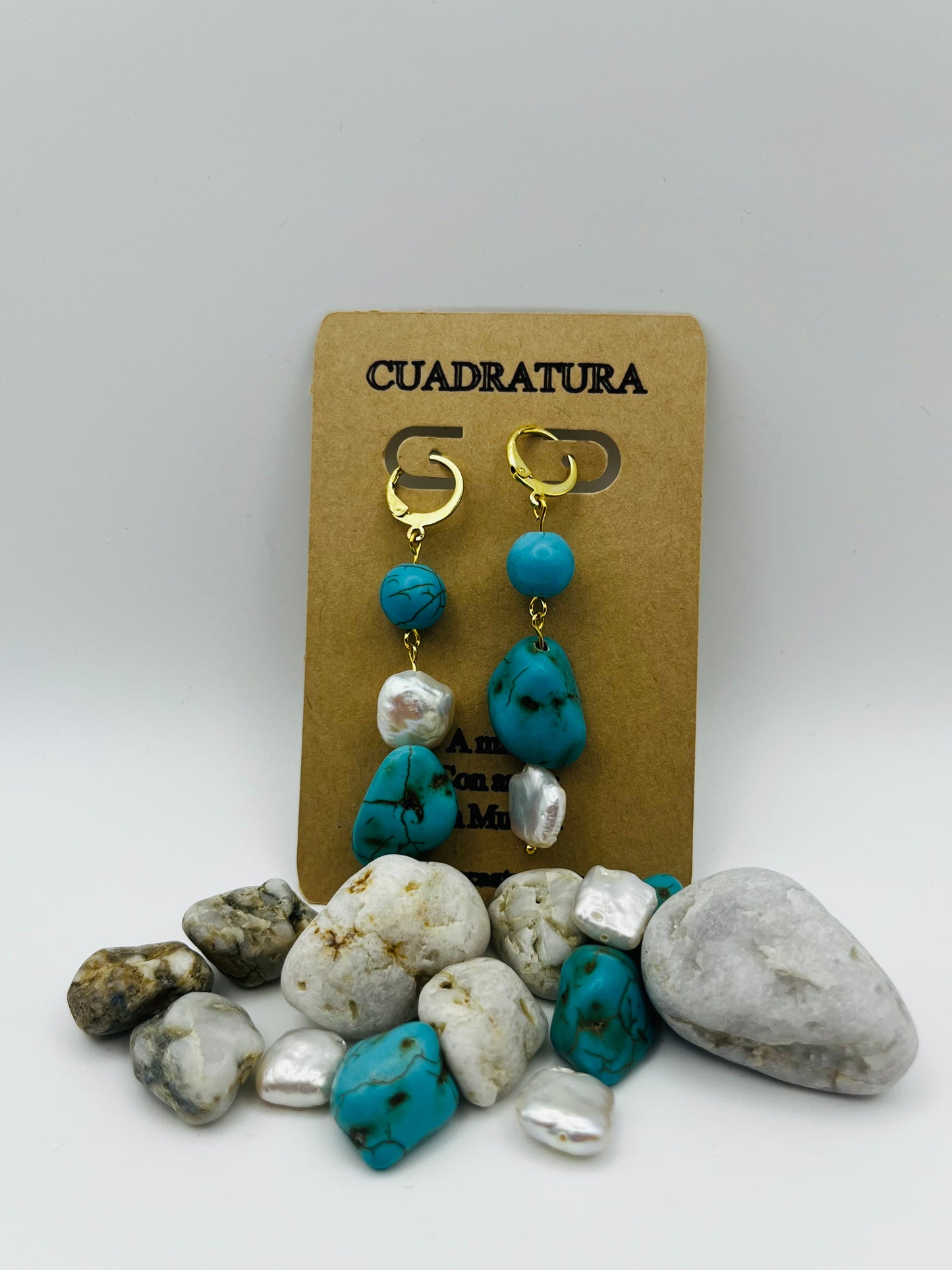 Pendientes Turquesa – pendientes asimétricos con aro dorado, cuenta redonda de howlita turquesa, perla keshi blanca y cuenta grande irregular de howlita turquesa con perla keshi en la base, en tarjeta Cuadratura, sobre montículo de piedras de playa blancas, cuentas de howlita turquesa y perlas keshi sueltas, fondo gris claro, hechos a mano en Murcia