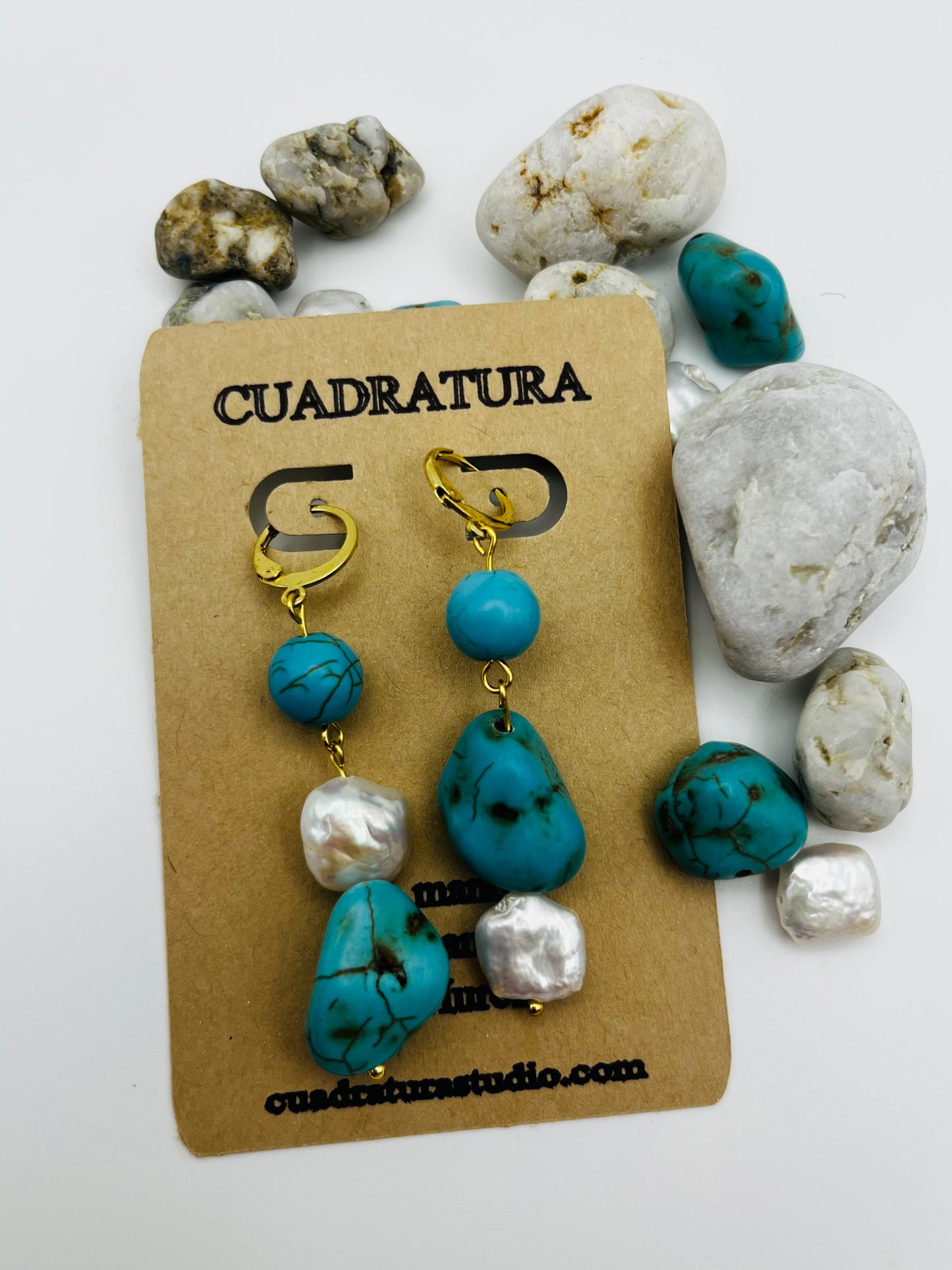Pendientes Turquesa – pendientes simétricos con aro dorado, cuenta redonda de howlita turquesa, perla keshi blanca y cuenta grande irregular de howlita turquesa con perla keshi en la base, en tarjeta Cuadratura tumbada, rodeada de piedras de playa blancas y cuentas de howlita turquesa sueltas, fondo blanco, hechos a mano en Murcia