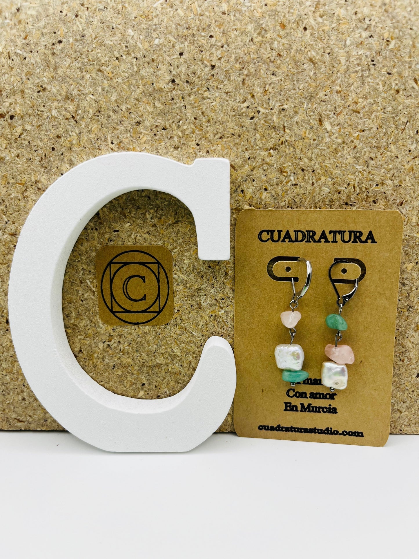 Pendientes Vera – pendientes simétricos con aro plateado, chip de cuarzo rosa, chip de amazonita verde menta y perla keshi blanca cuadrada, en tarjeta Cuadratura, sobre letra C blanca y fondo de corcho, hechos a mano en Murcia