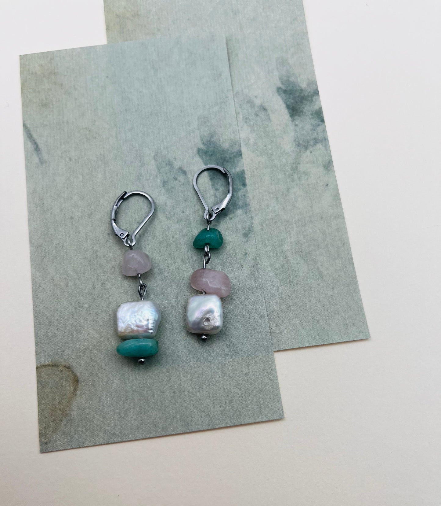 Pendientes Vera – dos aros plateados con chip de cuarzo rosa, chip de amazonita verde menta y perla keshi blanca cuadrada, desplegados sobre hojas de papel marmolado verde grisáceo superpuestas, fondo crema, hechos a mano en Murcia