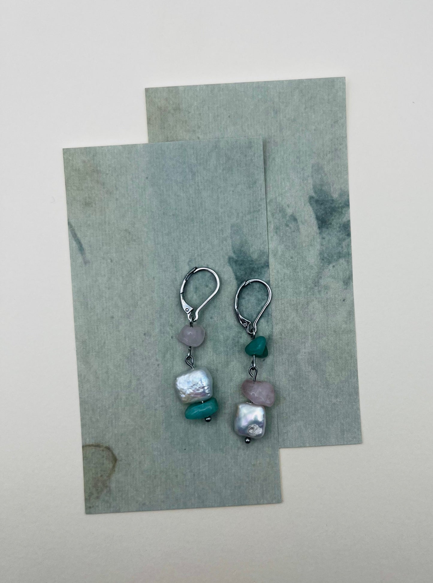 Pendientes Vera – dos aros plateados con chip de cuarzo rosa, chip de amazonita verde menta y perla keshi blanca cuadrada, sobre hojas de papel marmolado verde grisáceo superpuestas, fondo crema, hechos a mano en Murcia
