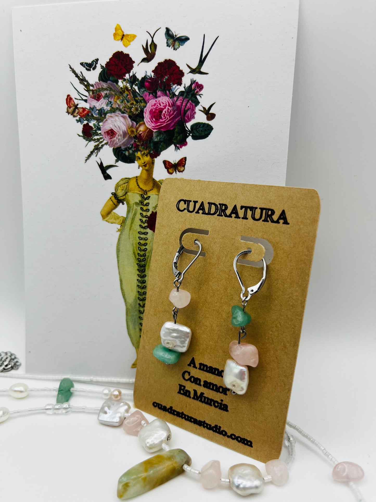 Pendientes Vera – pendientes simétricos con aro plateado, chip de cuarzo rosa, chip de amazonita verde menta y perla keshi blanca cuadrada, en tarjeta Cuadratura apoyada en postal ilustrada de dama con ramo de flores, rodeada de perlas keshi, chips de amazonita y cuarzo rosa sueltos, fondo blanco, hechos a mano en Murcia