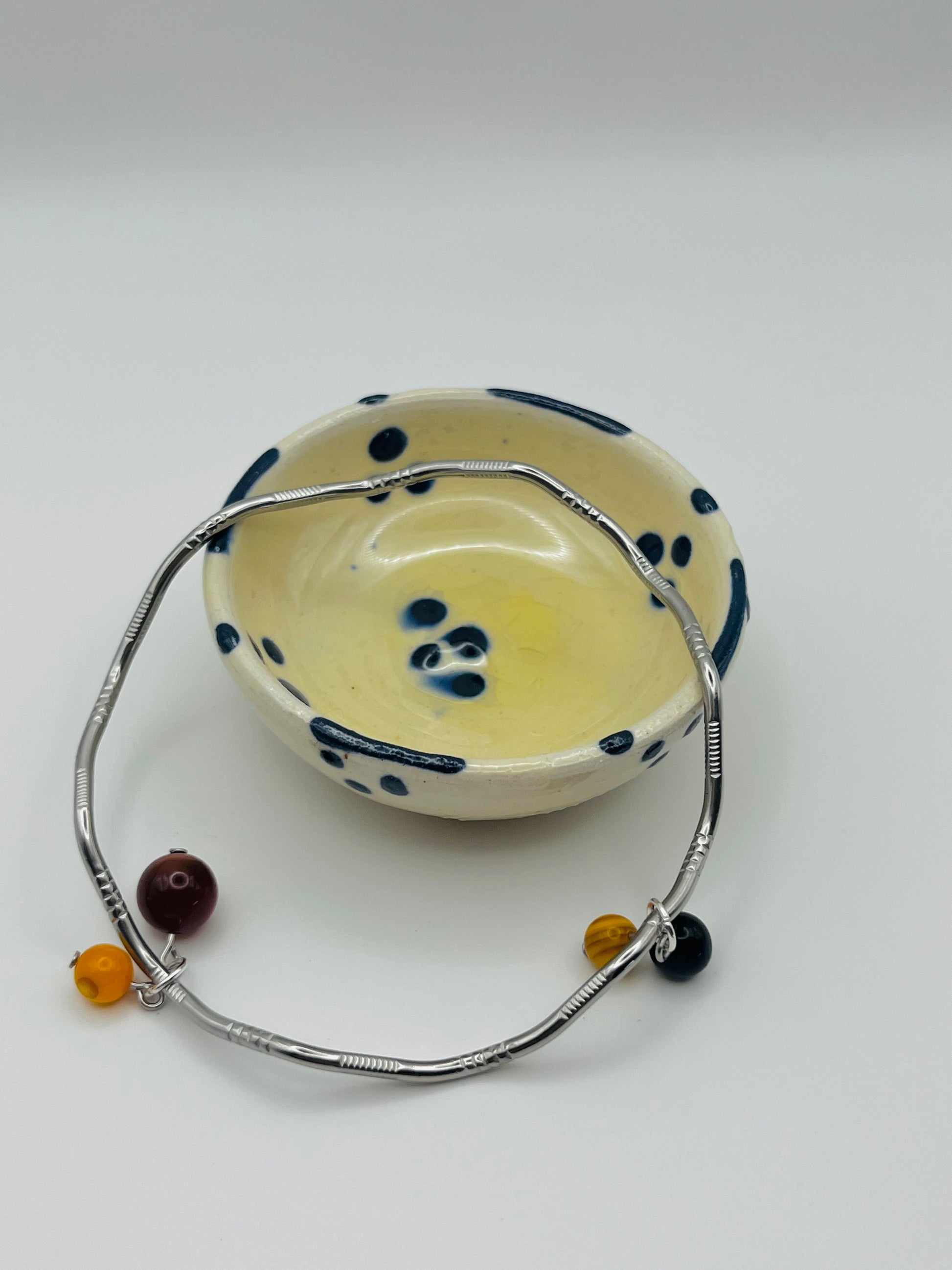 Pulsera artesanal Cala en aro rígido plateado con colgantes de lapislázuli azul, ágata roja y ágata amarilla, apoyada en cuenco de cerámica artesanal, montada a mano en Murcia
