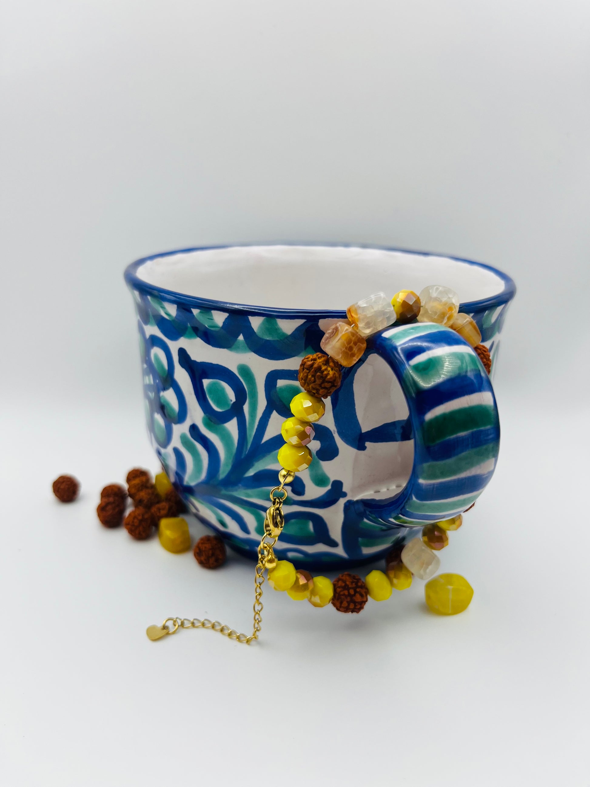 Pulsera artesanal Calima con semillas de rudraksha, cristal amarillo y nácar, colgada de taza de cerámica talavera azul, montada a mano en Murcia
